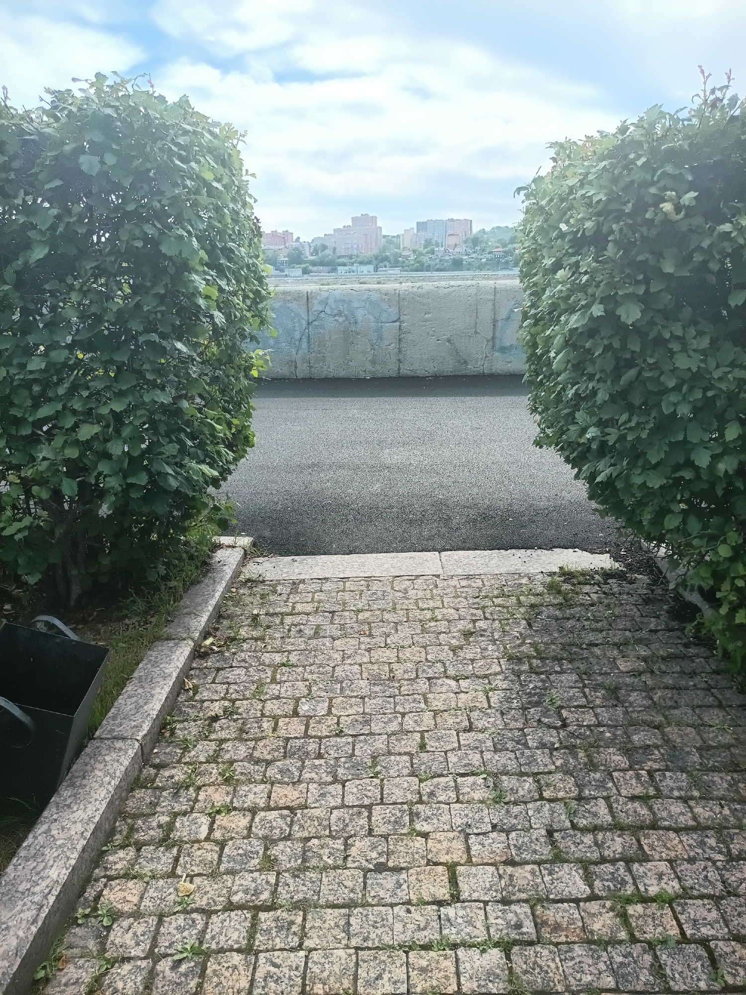 The image shows a pathway paved with small, irregularly shaped granite stones leading towards a darker asphalt surface. The pathway is bordered by lush green bushes on either side, framing the view ahead. Beyond the bushes, a grey concrete wall runs horizontally, with the asphalt surface directly in front of it. In the background, a cityscape of buildings is visible under a partly cloudy sky. The scene appears to be an outdoor public space, possibly a park or promenade. Without further information, it is impossible to determine the exact location within Irkutsk, Russia, or if there are any people or specific actions taking place.