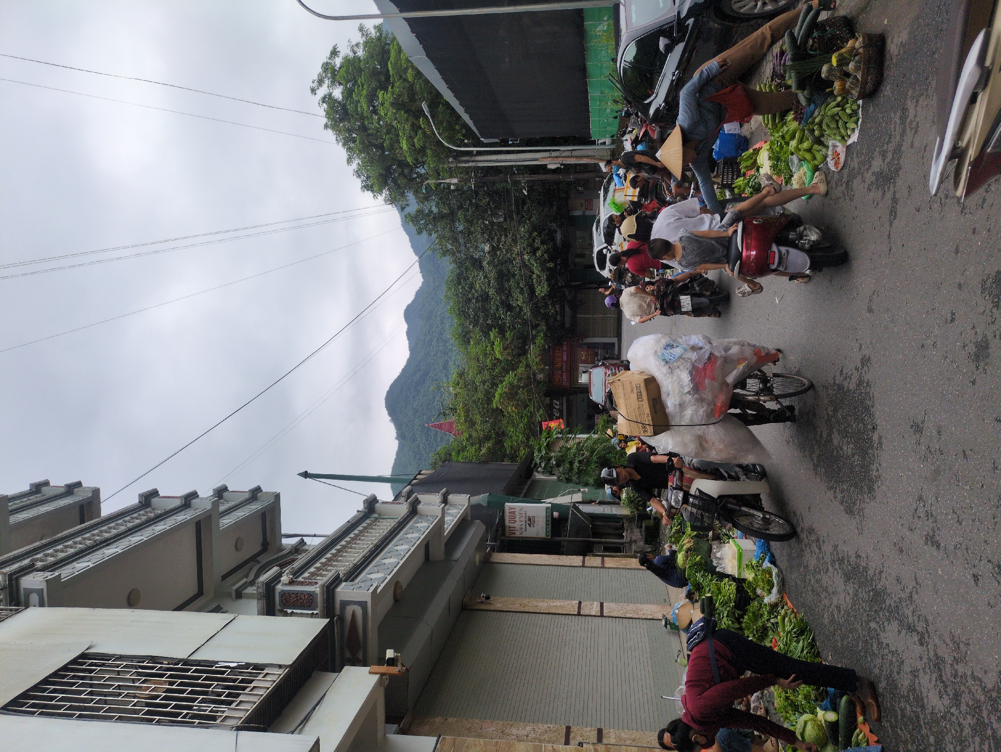 In Hà Giang 2 Ward, Vietnam, a street market unfolds under an overcast sky. A narrow street, wet from recent rain, is lined with buildings and a lush green hillside in the distance. People on motorcycles navigate the scene, some carrying large loads of goods. One man, wearing a dark shirt and helmet, rides a motorcycle loaded with a large, clear plastic bag filled with items. Another motorcycle is stopped, with a person in a conical hat attending to a roadside stall overflowing with fresh produce, including cucumbers, gourds, and other vegetables. Several individuals are seen browsing or arranging the produce. A woman in a maroon jacket is bent over a pile of vegetables, seemingly selecting items. A black car is parked on the right side of the street, adjacent to the produce stall. The general atmosphere suggests a busy, informal marketplace.