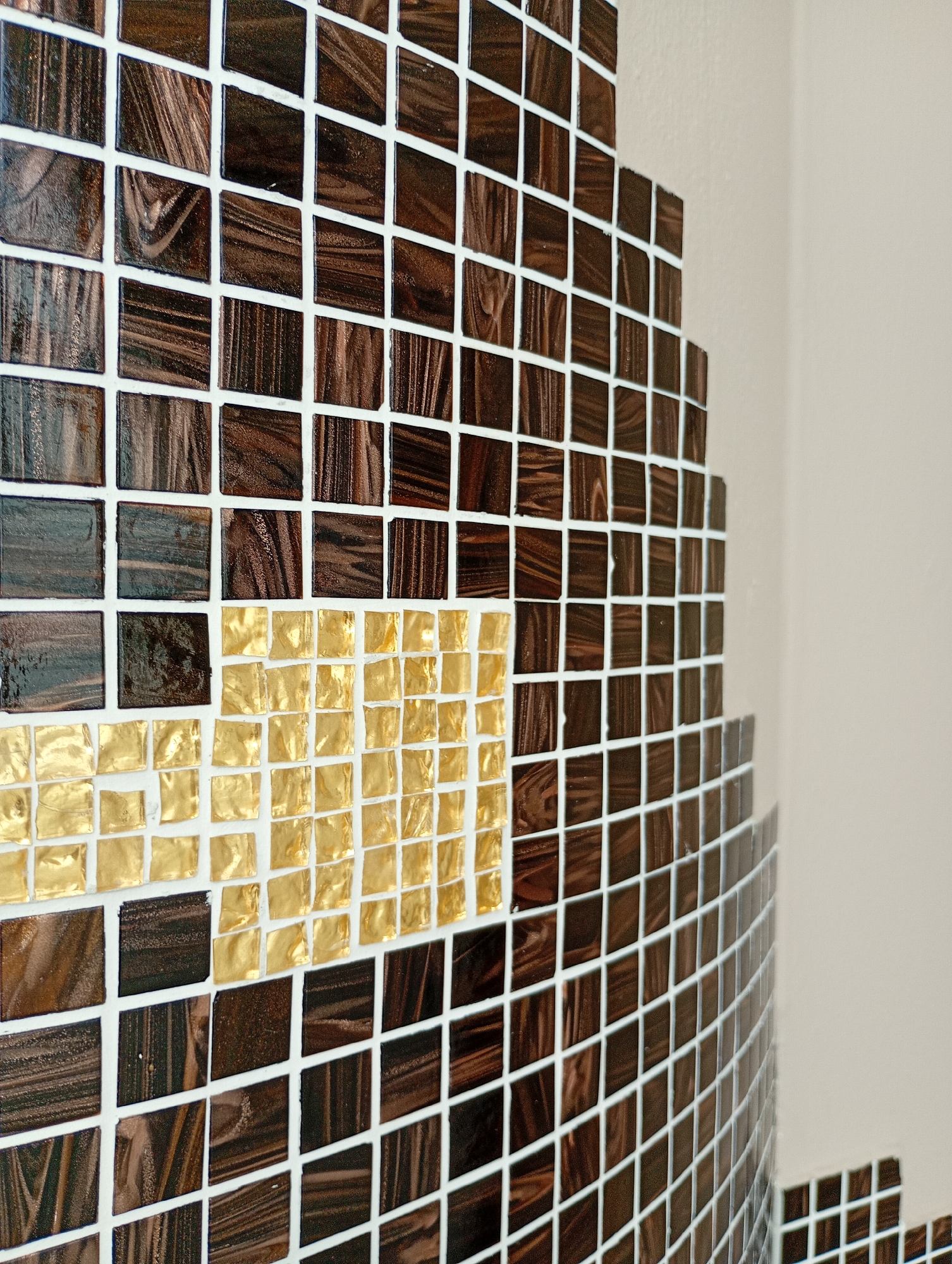 This image shows a close-up of a mosaic wall with small, square tiles. The majority of the tiles are a dark brown with a marbled, wood-like pattern and are arranged in a grid. A section of the mosaic features a cluster of small, square gold tiles, creating a contrasting accent. The mosaic appears to be on a curved surface, possibly a column or wall, and extends upwards and to the right. The background is a plain, light-colored wall. There are no people or animals visible in the image. The specific location in Bad Sachsa, Germany, cannot be determined from the image alone.