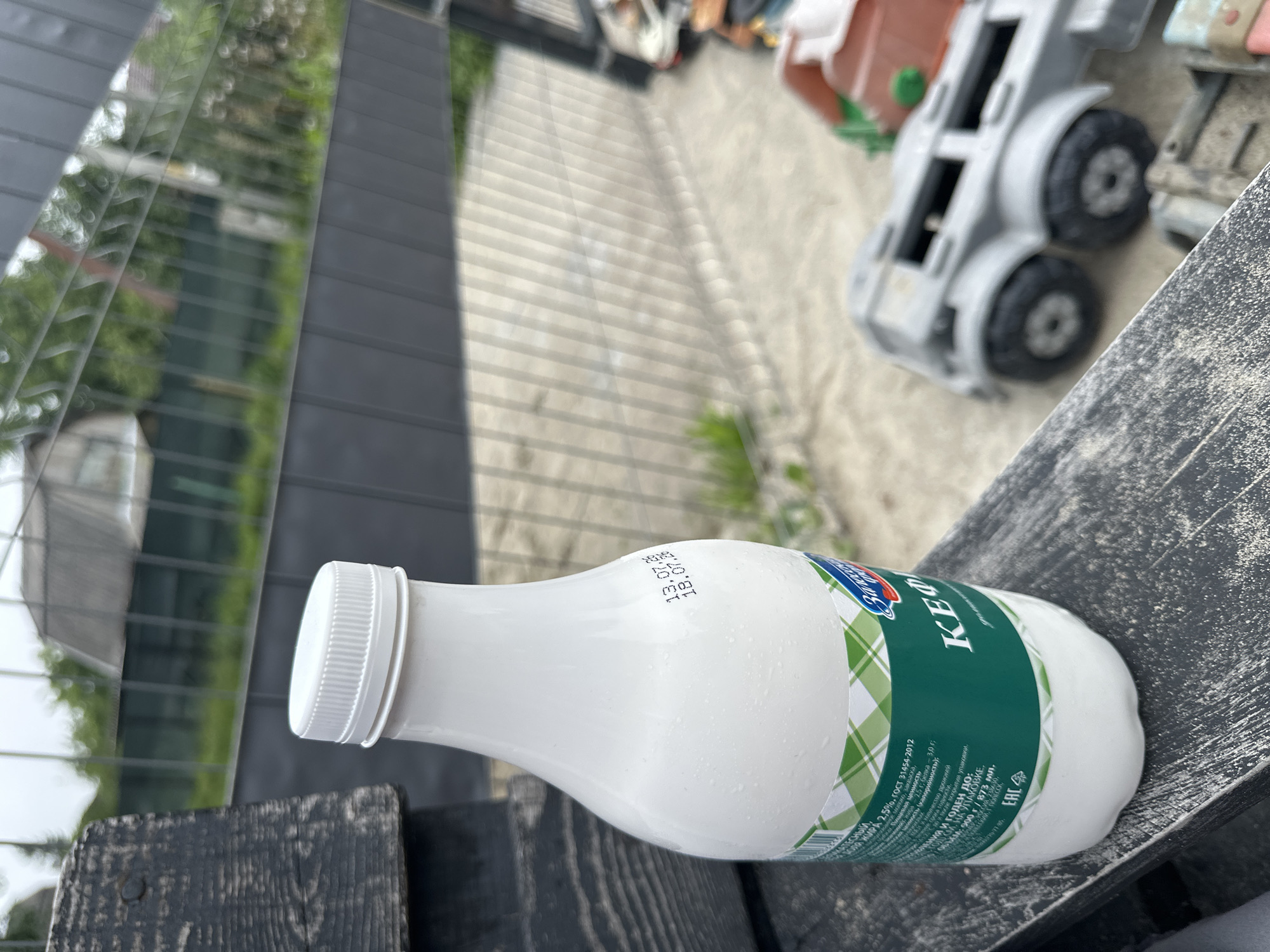 A close-up shot shows a white plastic bottle of "Кефир" (Kefir) with a green and white checkered label, resting on a dark wooden surface. The bottle has a white screw-on cap and some condensation on its exterior. The label features Cyrillic text and branding. Date stamps are visible on the side of the bottle, reading "13.07.26" and "18.07.26". In the background, a blurred scene of an outdoor environment is visible, including a grey metal fence, green foliage, and part of a building. To the right, out of focus, are several children's toys, including a grey toy truck and orange plastic construction equipment. The overall setting appears to be an outdoor space, possibly a backyard or patio. The location is specified as Malinovka, Russia.