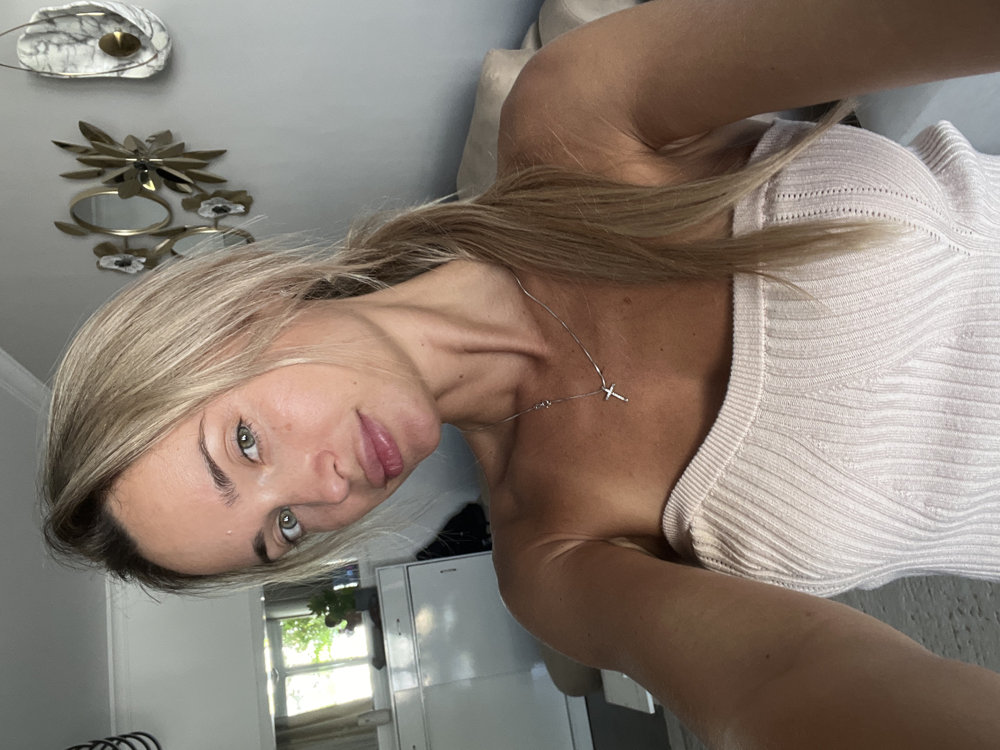 A woman with blonde hair pulled into a ponytail and green eyes is taking a selfie indoors. She is wearing a light pink, strapless ribbed top and a silver necklace with a cross pendant. Her skin appears tanned, and she has light makeup on. The background shows a room with a window, a white cabinet, a sofa, and wall decor. The scene is bright and appears to be daytime. There is no visible indication of the location being Sharm El Sheikh, Egypt.