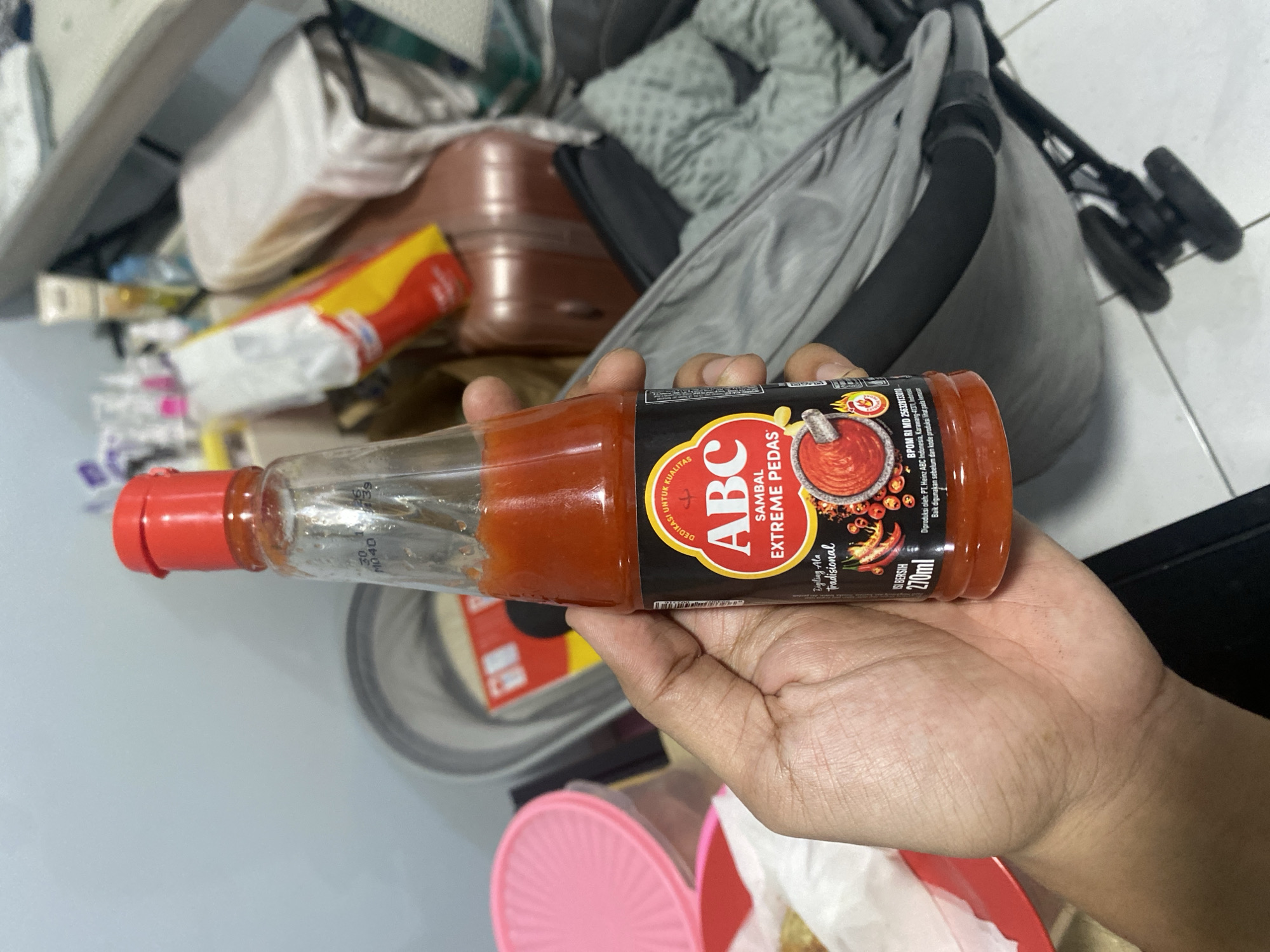 A hand holds a bottle of ABC Sambal Extreme Pedas sauce. The label on the bottle features the ABC logo in red and white, with the text "SAMBAL EXTREME PEDAS" below it. The sauce itself is red and appears to be a thick liquid. In the background, there are various objects, including what looks like a stroller, luggage, and some pink containers. The lighting suggests an indoor setting, possibly a home. There are no explicit geographical markers indicating the location in Dukuh, Indonesia, based solely on the visual information provided.