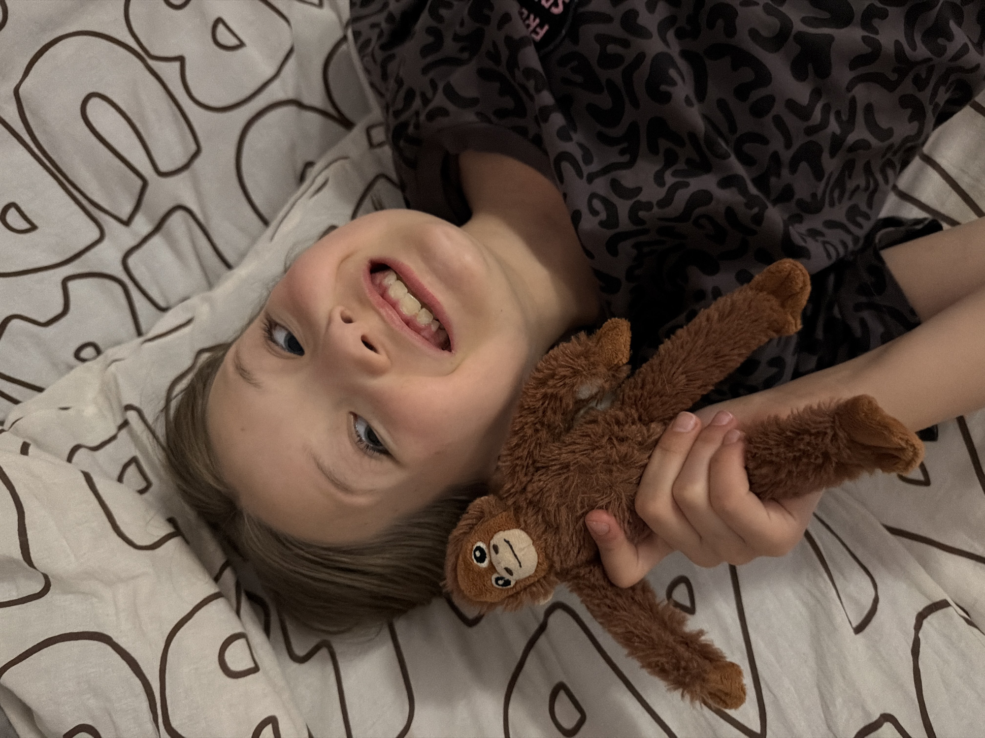 A young child is lying on a patterned bedsheet, smiling and holding a stuffed monkey. The child has blond hair and blue eyes. The bedsheet features a large, abstract pattern. The child is wearing a patterned top. The location is unknown.