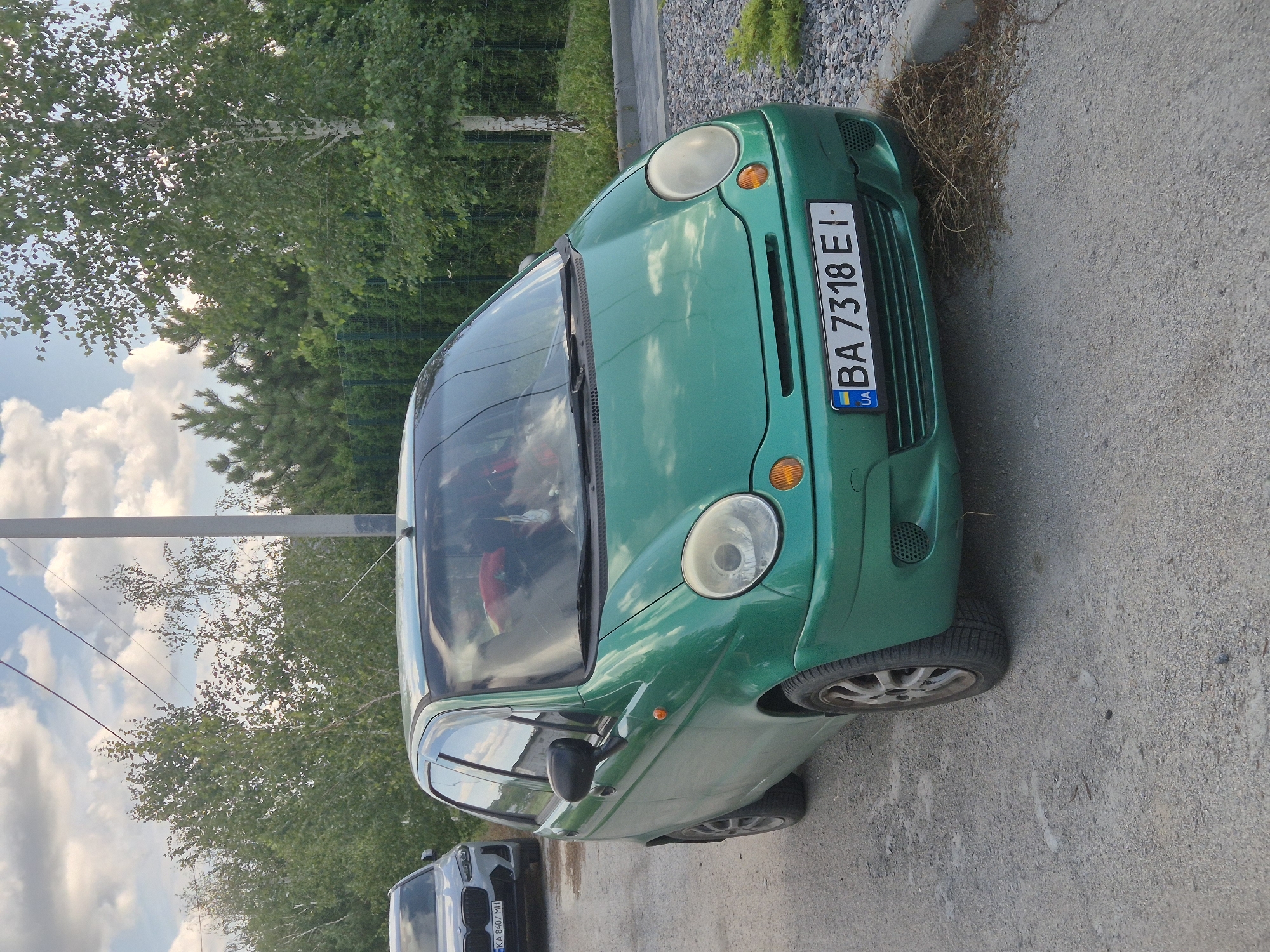 A green Daewoo Matiz is parked on a gravel surface next to a paved area. The car's license plate reads "BA 7318 EI" and has a Ukrainian flag emblem. In the background, there is a white BMW SUV, trees, a fence, and a utility pole. The sky is partly cloudy.