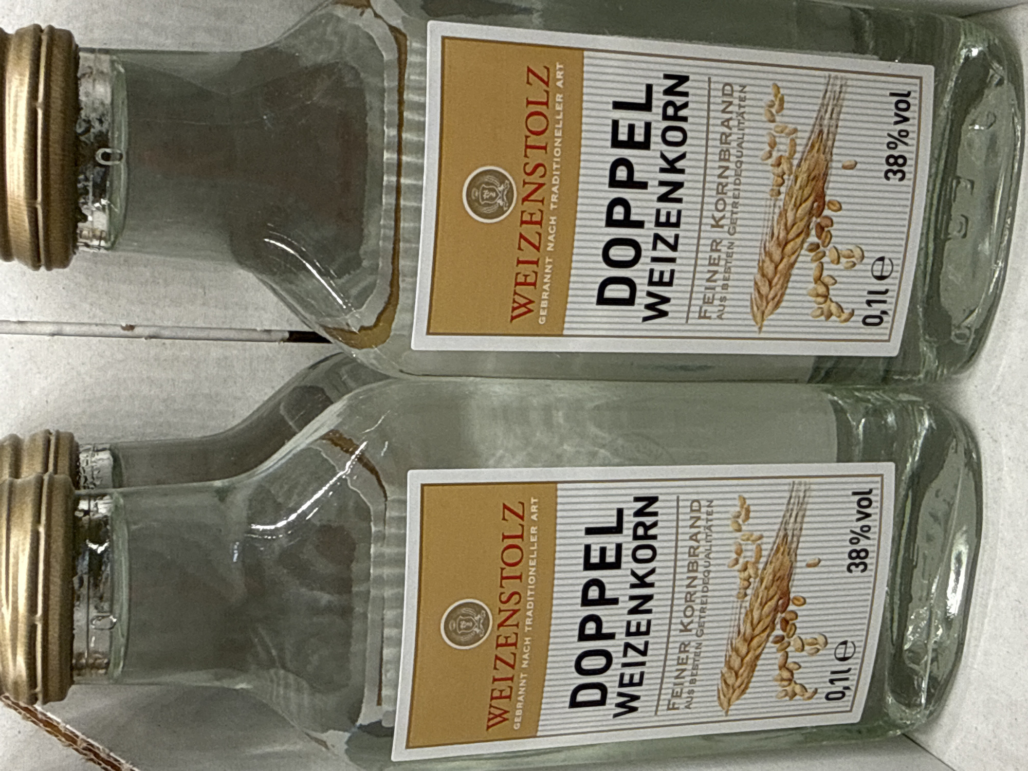 The image shows two small glass bottles of "DOPPEL WEIZENKORN" with golden caps, standing upright on a white surface. The bottles have labels that are predominantly gold and white, with red text. The labels read "WEIZENSTOLZ GEBRANNT NACH TRADITIONELLER ART DOPPEL WEIZENKORN FEINER KORNBRAND AUS BESTEN GETREIDEQUALITÄTEN". Below this text, there is an illustration of a wheat stalk and grains. At the bottom of the label, it states "0,1 le 38% vol". The bottles appear to be on display, possibly in a shop or a warehouse, given the white background and the presence of what looks like a cardboard divider between the bottles. The lighting is bright, casting subtle reflections on the glass bottles. There are no people visible in the image. The specific location in the city of Bad Lauterberg im Harz, Germany is not discernible from the provided image.