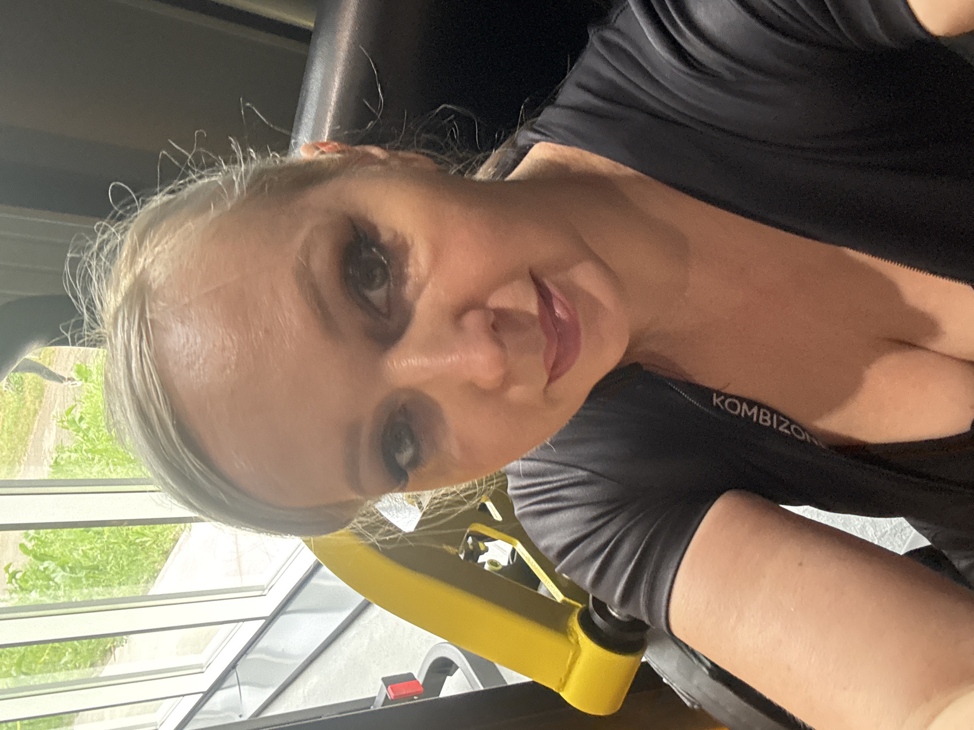 A blonde woman with makeup is taking a selfie in a gym. She is wearing a black top with the brand name "KOMBIAON" visible on the collar. The background shows a gym machine with yellow and black accents, as well as a window with green foliage outside. The woman is seated and looking directly at the camera. The scene appears to be indoors, within a fitness facility.