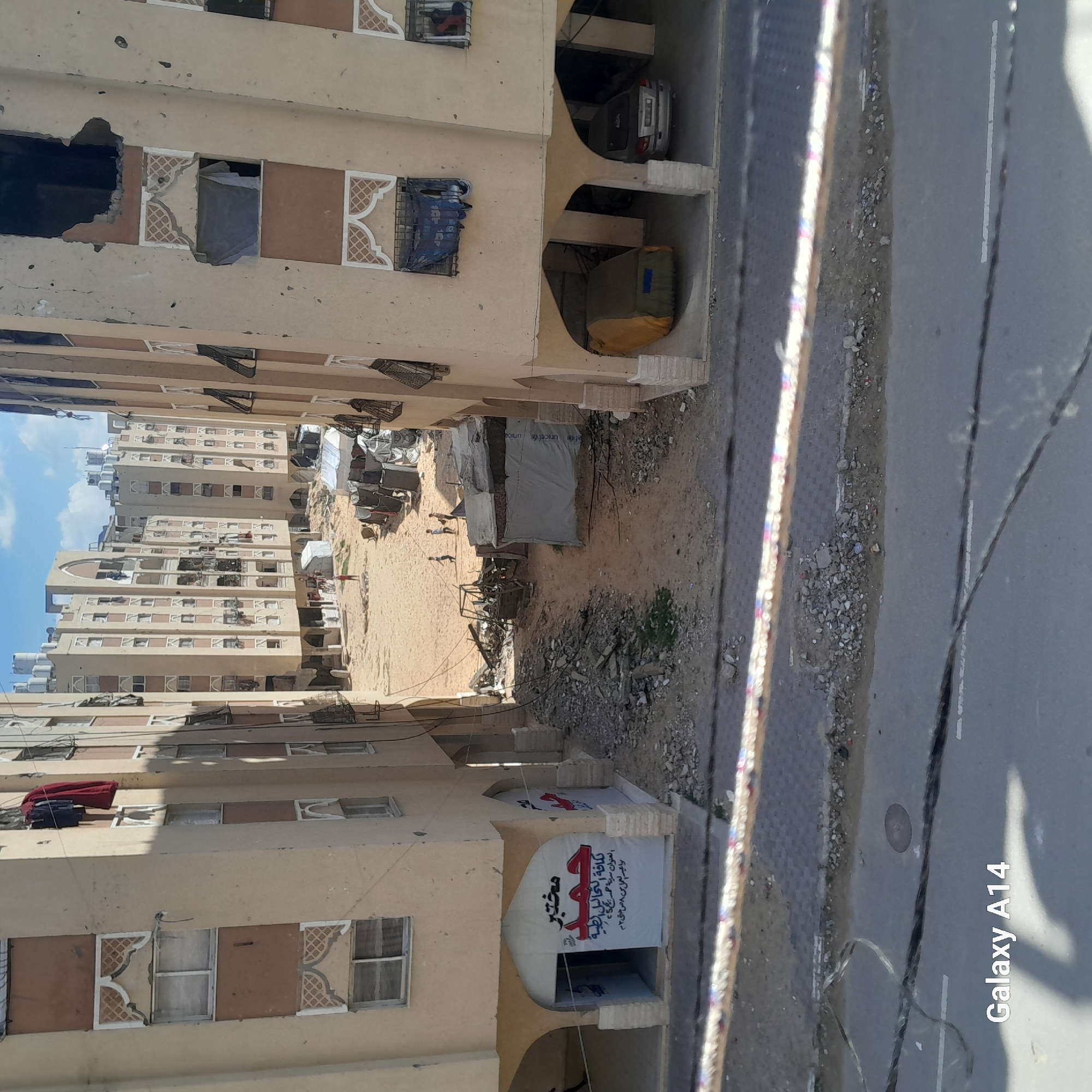 The image, taken in Khan Yunis, Palestinian Territory, shows a narrow alleyway between multi-story apartment buildings. The buildings are primarily light beige with patterned window frames. Some windows are damaged, with visible holes in the walls. In the alleyway, debris and rubble are scattered across the ground, and shelters marked with "UNICEF" can be seen. Several people are walking through the alley. A sign written in Arabic is visible near the bottom of the image. The sky is visible at the end of the alleyway, suggesting a clear day. A rope or wire is visible in the foreground. The camera's watermark "Galaxy A14" is in the bottom right corner.