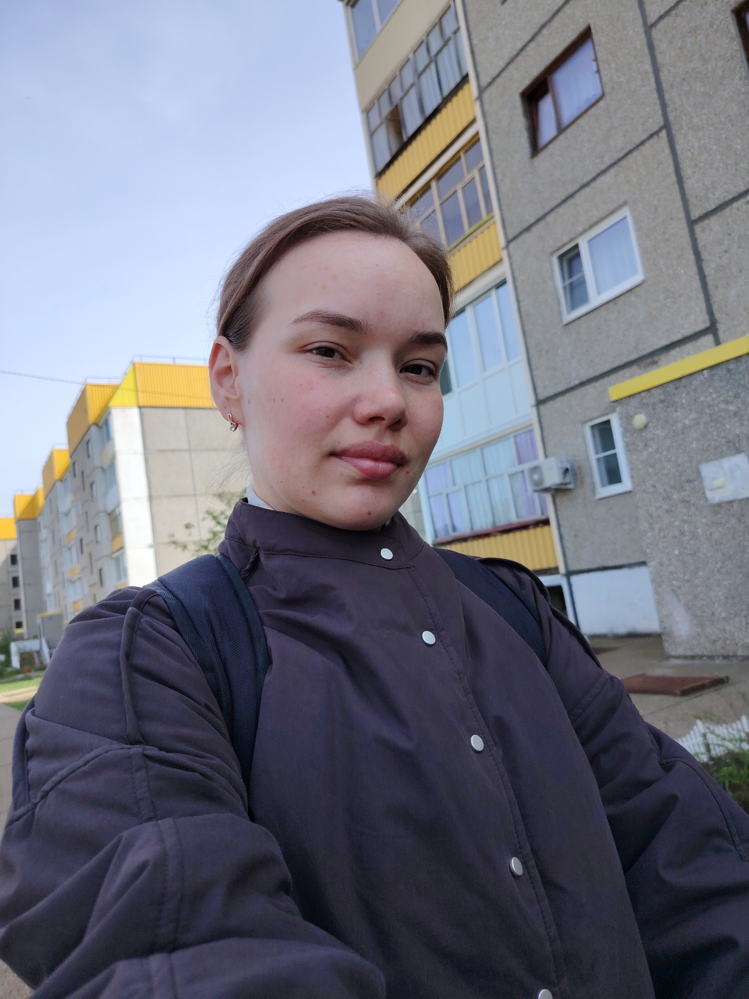 A young woman stands in front of a Soviet-era apartment building in Markovskii, Russia. She wears a dark-colored jacket with silver buttons and a backpack. The building is concrete, with yellow accents on the balconies and roofline. Several windows are visible, along with an air conditioning unit. The sky is light blue. The woman appears to be taking a selfie.