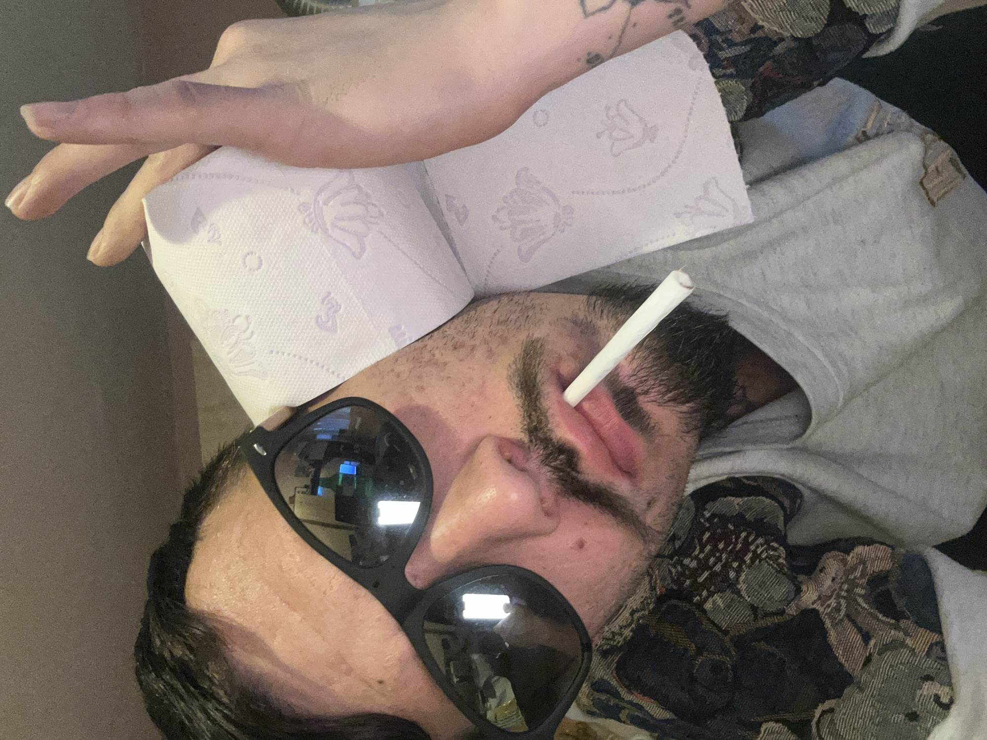 The image displays an adult male in an indoor setting, likely a home, captured from a close-up, slightly tilted perspective. He is lying down, with his head resting against an indistinct, dark background. The photo is rotated, making the subject appear to be on his right side.
The man wears black sunglasses that reflect bright, artificial light sources, possibly from a computer screen, showing blue and white elements. A white, cylindrical object, resembling a cigarette or vape pen, is positioned between his lips. A roll of white toilet paper, adorned with light purple butterfly and floral patterns, is resting on his head or in his hair.
He is dressed casually in a light grey hooded sweatshirt. Around his neck and shoulder, a section of a dark, intricately patterned fabric, possibly a blanket or part of another garment, is visible. Faint, dark tattoos can be seen on what appears to be his bare arm or shoulder, partially visible beneath the toilet paper. His face features stubble or a short beard, and there is a noticeable reddish area or irritation near his left eye (viewer's right side).
The overall impression is one of casual relaxation or repose. The reflections in his sunglasses suggest artificial lighting, making the time of day ambiguous. No specific visual cues within the image confirm the location as Varna, Bulgaria. Beyond the distinct butterfly and floral patterns on the toilet paper, no discernible text is visible in the image.