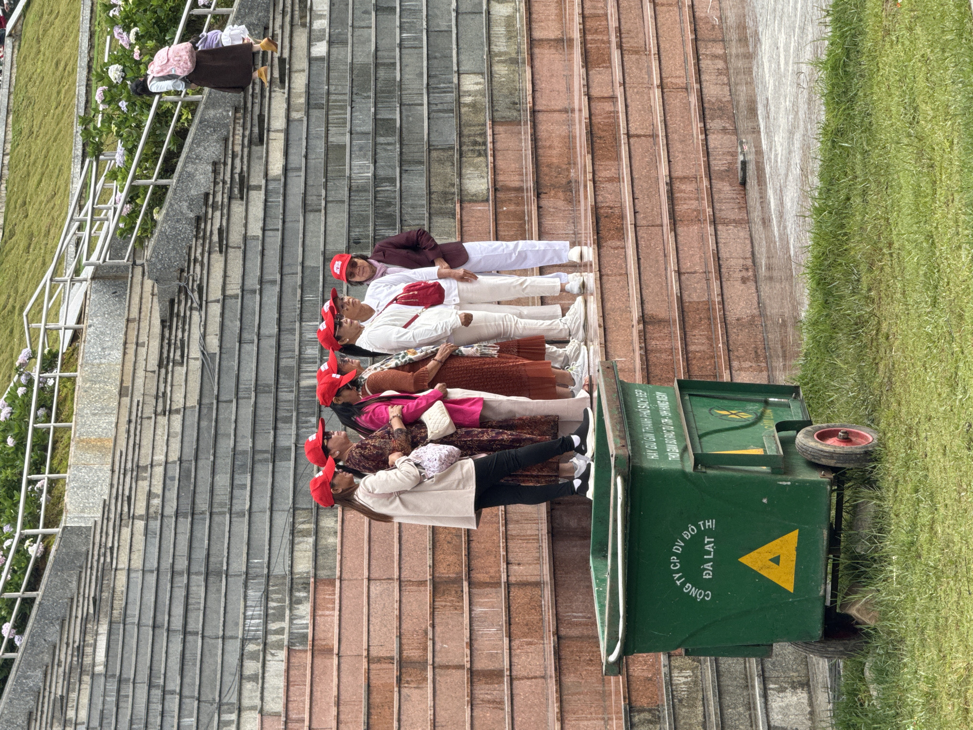 An outdoor scene in Da Lat, Vietnam, depicts a group of people positioned on a series of steps next to a green area. When viewed upright, the image features multiple sets of stairs, with grey concrete steps on the left and reddish-brown tiled steps on the right. A lush green lawn borders the left side of the stairs, accompanied by bushes with light-colored flowers and metal handrails.
A group of six individuals, predominantly women, is lined up on the reddish-brown steps in the mid-ground. All are wearing identical red baseball caps. Their clothing varies, including white tops, a maroon jacket, and a mix of light and dark pants, with some carrying small bags. They appear to be posing or waiting, looking towards the left side of the frame. Further up the grey stairs, another person with a pink backpack and a brown skirt is visible.
In the foreground, on the right, a large green municipal waste bin with wheels is prominent. The bin has yellow text and a triangular symbol, and includes Vietnamese phrases: "CÔNG TY CP DV ĐÔ THỊ ĐÀ LẠT" (Da Lat Urban Services Joint Stock Company), "HÃY GIỮ GÌN THÀNH PHỐ SẠCH ĐẸP" (Please keep the city clean and beautiful), and "THỜI GIAN ĐỔ RÁC TỪ 17H-19H HÀNG NGÀY" (Garbage collection time from 5 PM - 7 PM daily). The scene takes place during the daytime, under what appears to be overcast or diffused light, and suggests a public area like a park or garden in Da Lat.