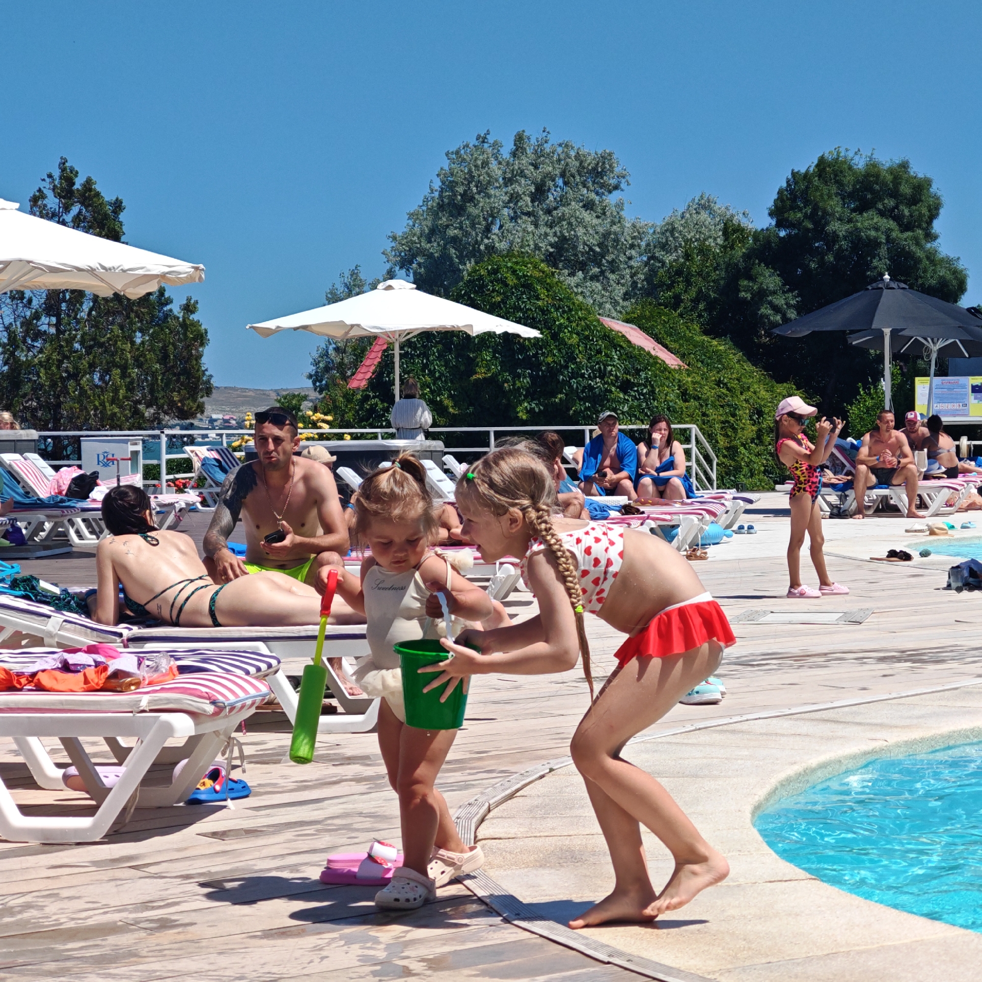 In a sun-drenched outdoor setting, likely a resort or public pool area, a group of people are relaxing on a wooden deck. The focus is on two young girls playing near the edge of a bright blue swimming pool. The girl on the left, wearing a light-colored sleeveless top and a white skirt, is holding a green bucket and a green toy water squirter. The girl on the right, in a red and white polka dot swimsuit with a red ruffled skirt, is looking towards the other girl, with her arms outstretched as if reaching for something. In the background, several adults are seated or reclining on lounge chairs, some under white umbrellas. A man with tattoos is sitting up on a lounge chair, holding a phone. Further back, other people are visible, including a woman in a blue robe. The environment is characterized by lush green trees and a clear blue sky, suggesting a warm, pleasant day. The wooden deck appears damp in places, indicating recent activity or proximity to the pool.