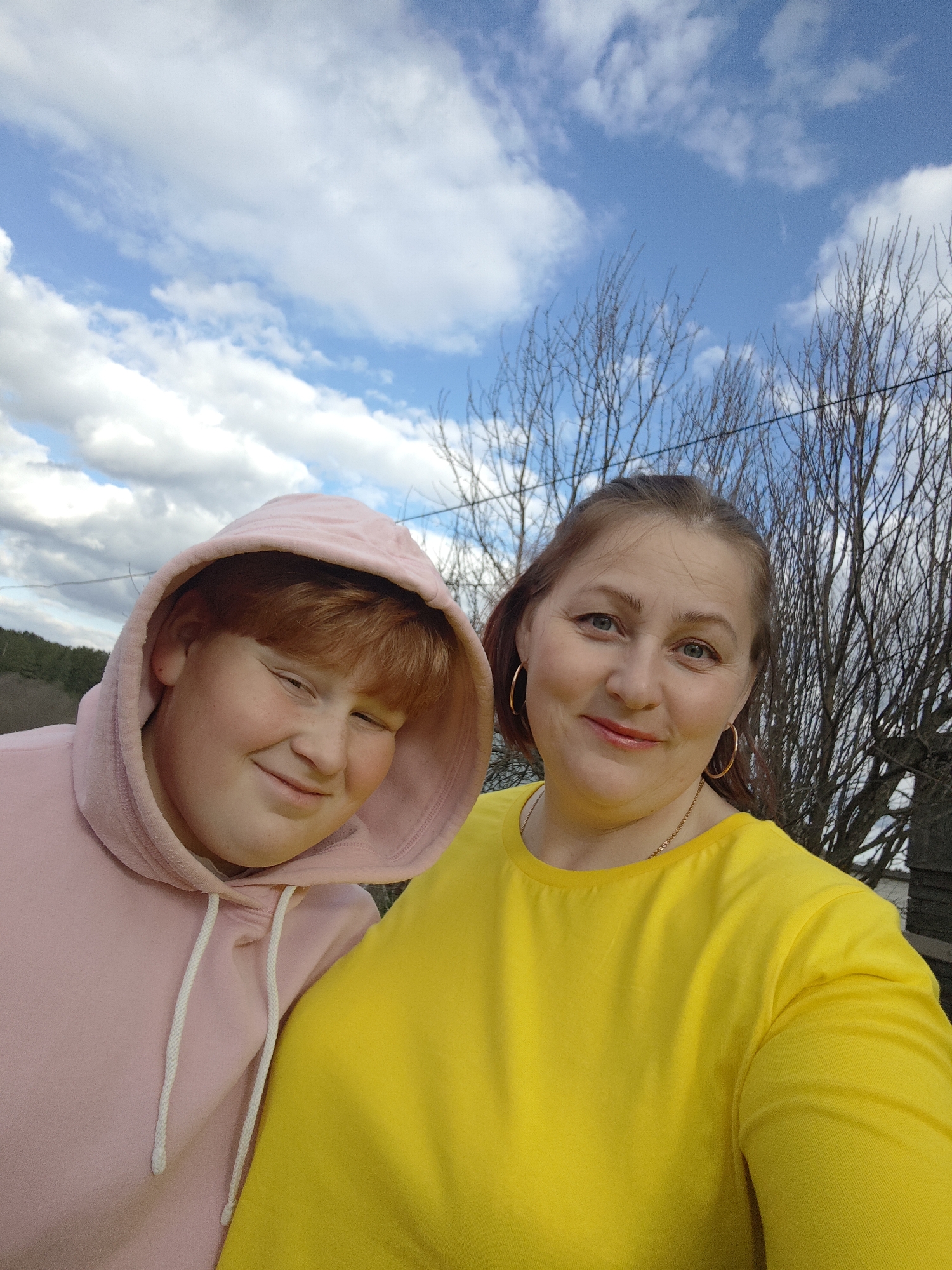 An outdoor selfie features an adult woman and a younger person, both smiling at the camera. The woman, positioned on the right, has light brown to auburn hair, light-colored eyes, and is wearing a bright yellow long-sleeved top, gold hoop earrings, and a thin gold chain. She has light makeup, including lipstick. To her left, the younger person, likely a boy, has red hair and freckles, wearing a pink hooded sweatshirt with the hood pulled up. He is also smiling broadly, with his eyes squinted.
The background showcases a clear blue sky dotted with white, fluffy clouds. Bare tree branches, characteristic of late winter or early spring, are visible behind both subjects. In the distance to the left, a forested hillside can be seen. The scene is brightly lit by natural daylight. Both individuals appear cheerful. No text is visible in the image. The setting is in Фоки, Russia.