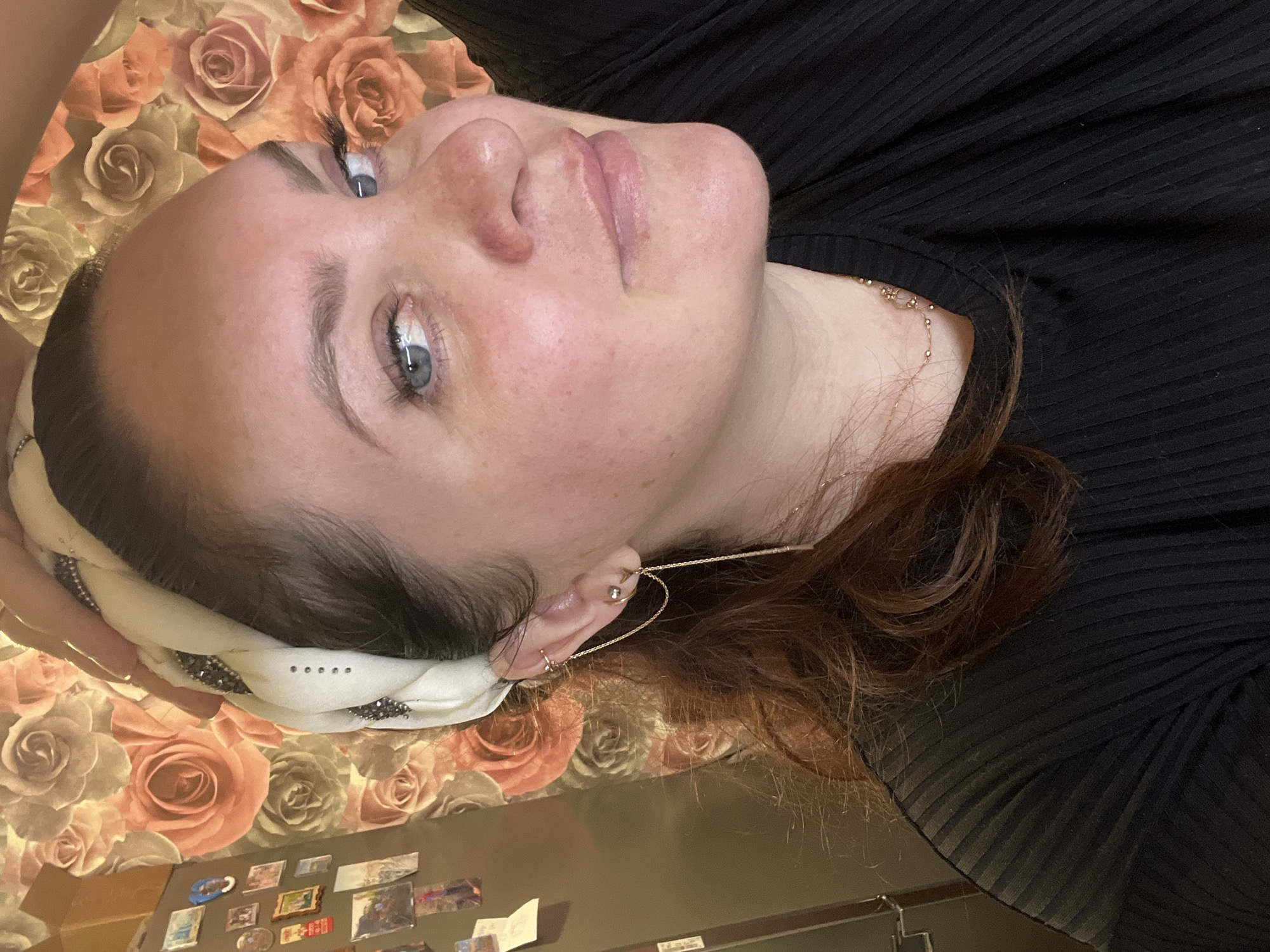 A woman with fair skin and light blue eyes is lying on a surface patterned with roses, her head tilted back. She is wearing a white headband with dark embellishments and a black shirt. Her reddish-brown hair is visible near her ear, which is adorned with several piercings and a long, delicate gold earring. A gold necklace is also visible around her neck. The bottom of the frame shows a surface with various small items, potentially magnets, attached to it. There is no indication of the city of Noviy, Russia, in the scene.