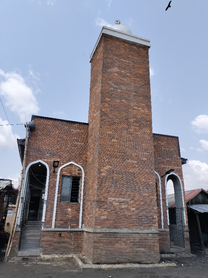 The image displays a mosque constructed primarily of reddish-brown brick, situated in Oworonshoki, Nigeria, under a partly cloudy sky. The building features a prominent, rectangular minaret on the right, topped with a light-colored dome and a golden crescent moon and star finial. A dark bird is visible flying in the upper right quadrant of the sky.
The main structure of the mosque has a dark roofline and multiple arched openings framed with white trim. On the left side of the building, there are two distinct arched openings. The leftmost opening functions as an entrance with several steps leading up to it, featuring metal grating at the base and a dark interior visible. Above this entrance, a grey horn speaker is mounted on the wall, partially obscured by power lines and wires. Adjacent to this entrance is another arched opening containing a window with horizontal security bars and a dark, possibly reflective, pane. A black rectangular light fixture is mounted on the brick wall between these two arches.
The ground directly in front of the mosque appears to be dirt or unpaved, with a raised concrete curb segment separating it from the building's base. To the far left and right of the mosque, rudimentary structures with corrugated metal roofs and weathered facades are partially visible, suggesting a dense urban or semi-urban setting. No individuals or direct interactions are visible in the scene.