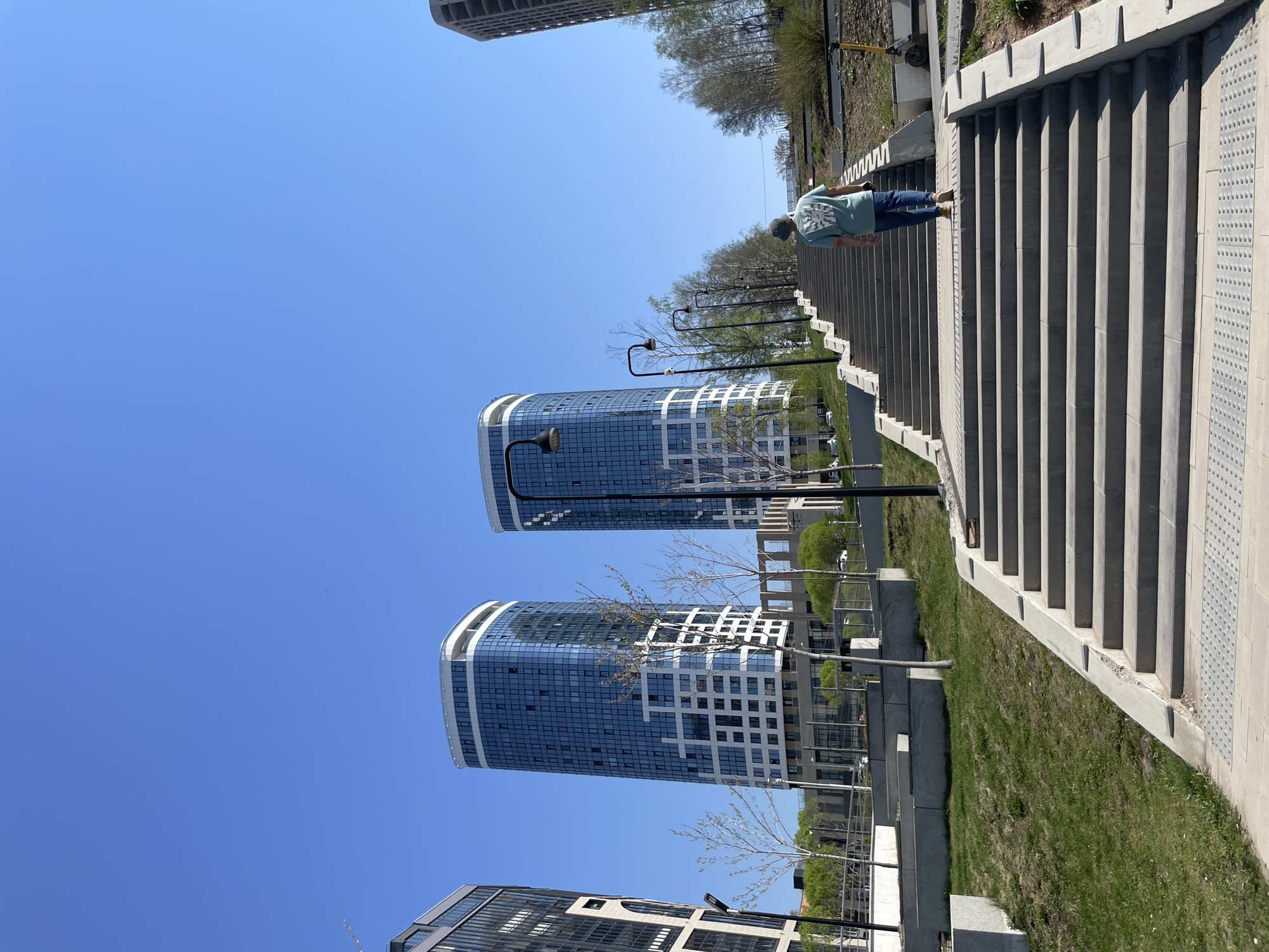 The image shows an outdoor scene, with a person walking up a long set of concrete stairs. The person is wearing a green shirt, blue jeans, and a cap, and is bending forward slightly. Parallel to the stairs is a strip of green grass and several modern buildings with glass facades. Street lamps are spaced along the side of the stairs. The sky above is clear blue. According to web searches, the location is Izhevsk, Russia.