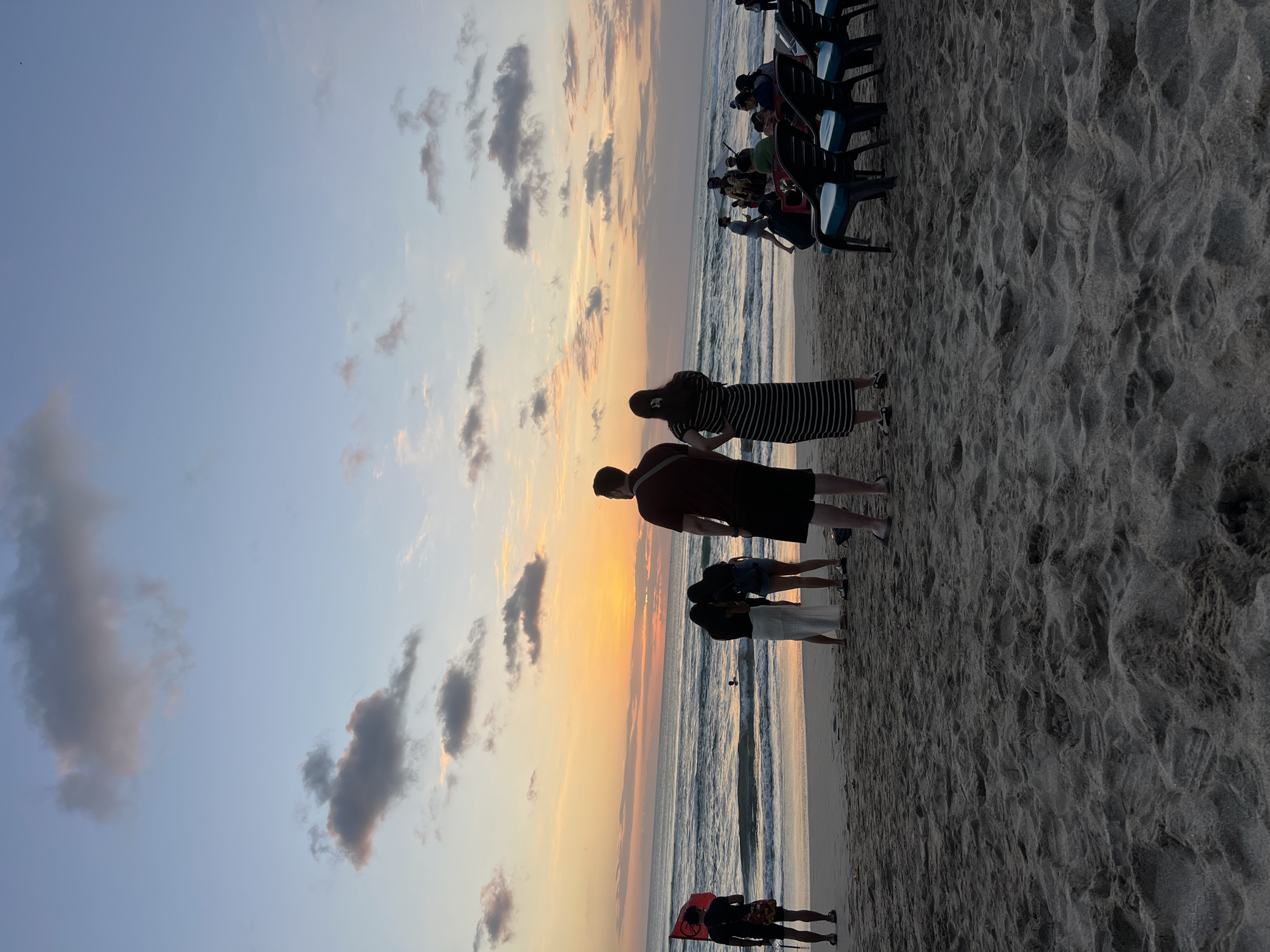 This image depicts a beach scene in Kuta, Indonesia, at sunset. The sky is a gradient of orange and blue, with scattered clouds. The sun is low on the horizon, casting a warm glow over the ocean and the sandy beach.
In the foreground, a sandy beach is visible, with footprints and some scattered debris. Several people are present on the beach. On the left, a person in a black shirt and shorts stands with a red and white flag. To the right of this person, a group of three people are standing together, with two women looking down and a man in a maroon shirt and black shorts standing with his back to the camera. Further to the right, a group of people are seated in chairs, facing the ocean.
The ocean is calm, with gentle waves rolling onto the shore. The water is a deep blue, reflecting the colors of the sunset. In the distance, the horizon line is visible, separating the ocean from the sky. The overall atmosphere of the image is serene and peaceful, capturing the beauty of a tropical sunset.