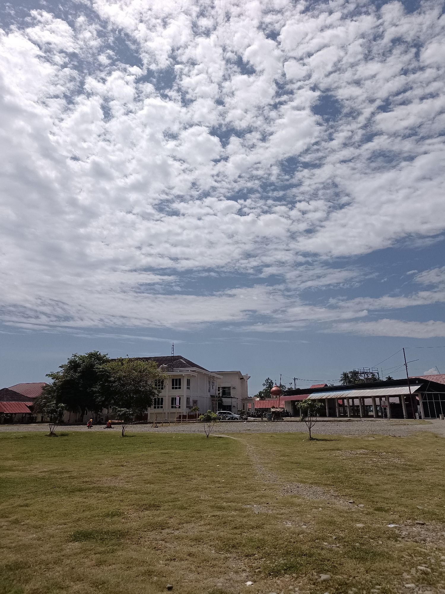 The image depicts an outdoor scene with a building complex and a grassy field under a cloudy sky. The building in the center is a multi-story white structure with numerous windows and a dark roof. To the left of this building, another structure with a reddish roof is partially visible. To the right, a long, covered open-air building with a white roof supported by columns is present, and behind it, another building is visible. Several trees are scattered throughout the scene, with a large cluster of trees behind and to the left of the central white building. A paved or gravel area is visible in front of the buildings. The foreground is dominated by a large expanse of dry, green grass. Two small figures, possibly people, are visible in the middle ground on the grass near the trees and the gravel area. One figure appears to be wearing red and the other is wearing yellow, suggesting they might be construction workers or engaged in some outdoor activity. The sky is filled with a mix of white cumulus clouds and patches of blue sky. The overall impression is of a developed area, possibly a school, institution, or commercial complex, situated in a location with open grounds. Given the context provided about the city of Mesjid Reuntoh, Indonesia, this scene could be a facility within that region.