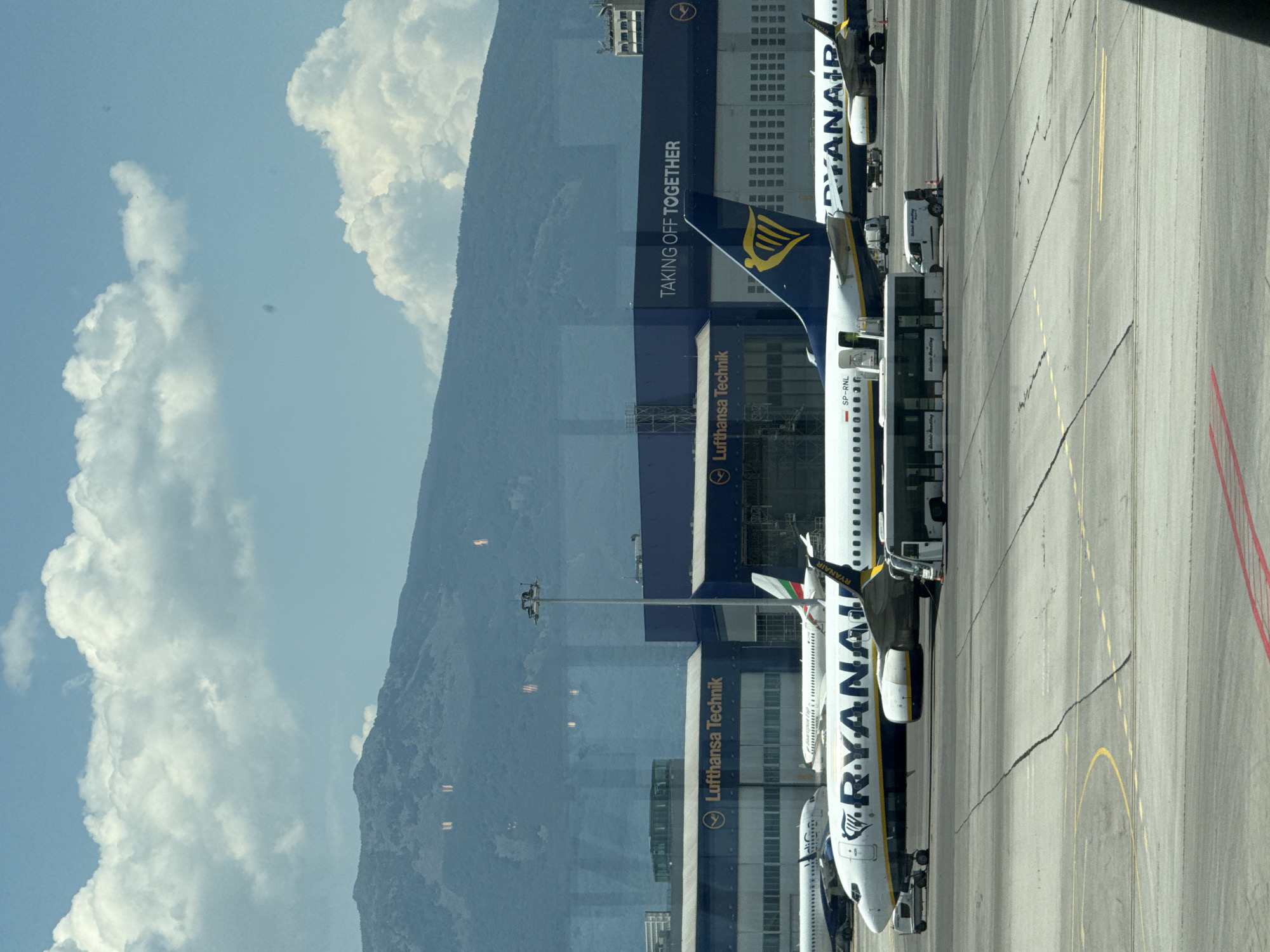 The image captures a scene at Sofia Airport in Bulgaria. In the foreground, an airplane with "RYANAIR" branding on its fuselage is parked on the tarmac. A jet bridge is connected to the aircraft, and a bus is parked beside it. In the background, another airplane with a Bulgarian flag on its tail is visible. Several hangars with the "Lufthansa Technik" logo are also present, along with a building that reads "TAKING OFF TOGETHER." Mountains form the backdrop under a sky filled with clouds. The overall environment suggests a functional airport setting on a day with partly cloudy skies.