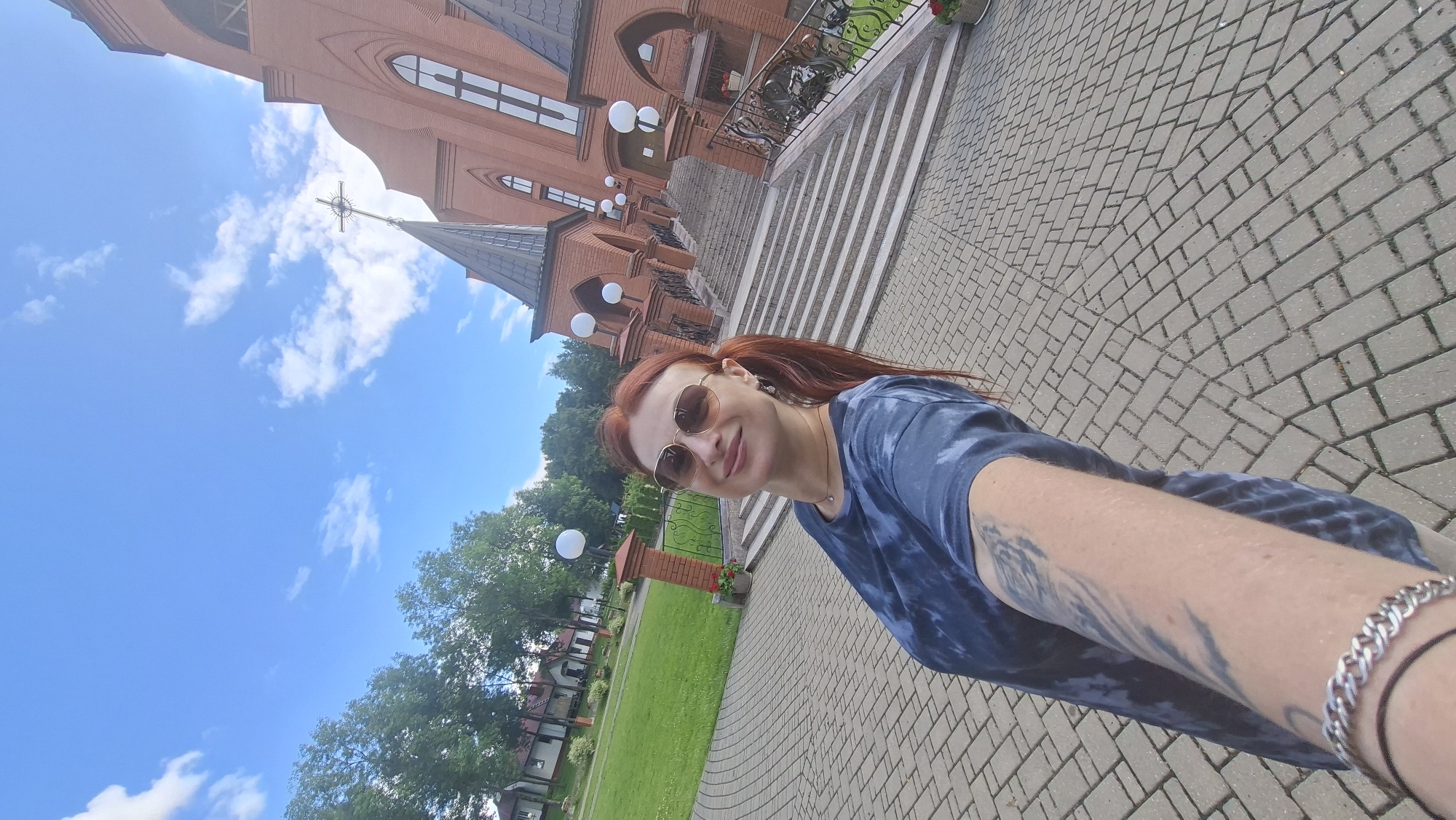 A woman with red hair pulled into a ponytail and wearing sunglasses and a dark blue tie-dye t-shirt is taking a selfie outside a brick church with a tall steeple and a cross. The church has a long set of stone stairs leading up to its entrance, with ornate railings and lampposts. The ground is paved with interlocking bricks. In the background, there is a green lawn, trees, and some buildings. The sky is blue with scattered white clouds.