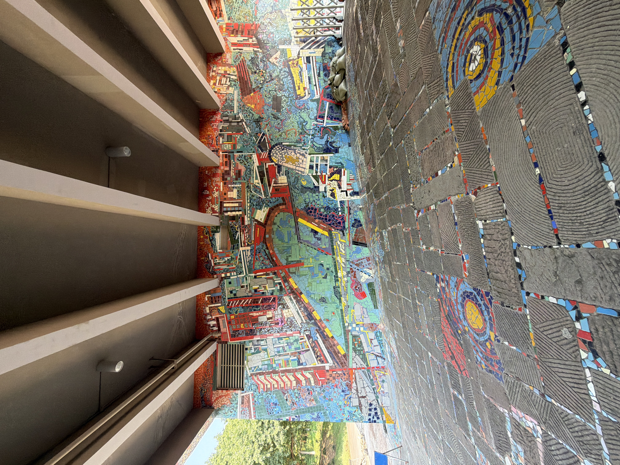 The image depicts a colorful mosaic mural on a wall and a mosaic-tiled floor beneath a concrete overpass in the Chongqing High-tech Zone, China. The mural features an abstract, city-like design with vibrant colors and geometric shapes, representing buildings, roads, and various urban elements. The floor is paved with large, textured gray tiles interspersed with smaller, colorful mosaic tiles forming circular and linear patterns. In the right background, several sandbags are piled up next to a metal fence, and two individuals are resting or sleeping on the mosaic floor, partially obscured by the sandbags. The scene is illuminated by natural light coming from the left and overhead lighting fixtures attached to the underside of the overpass.
