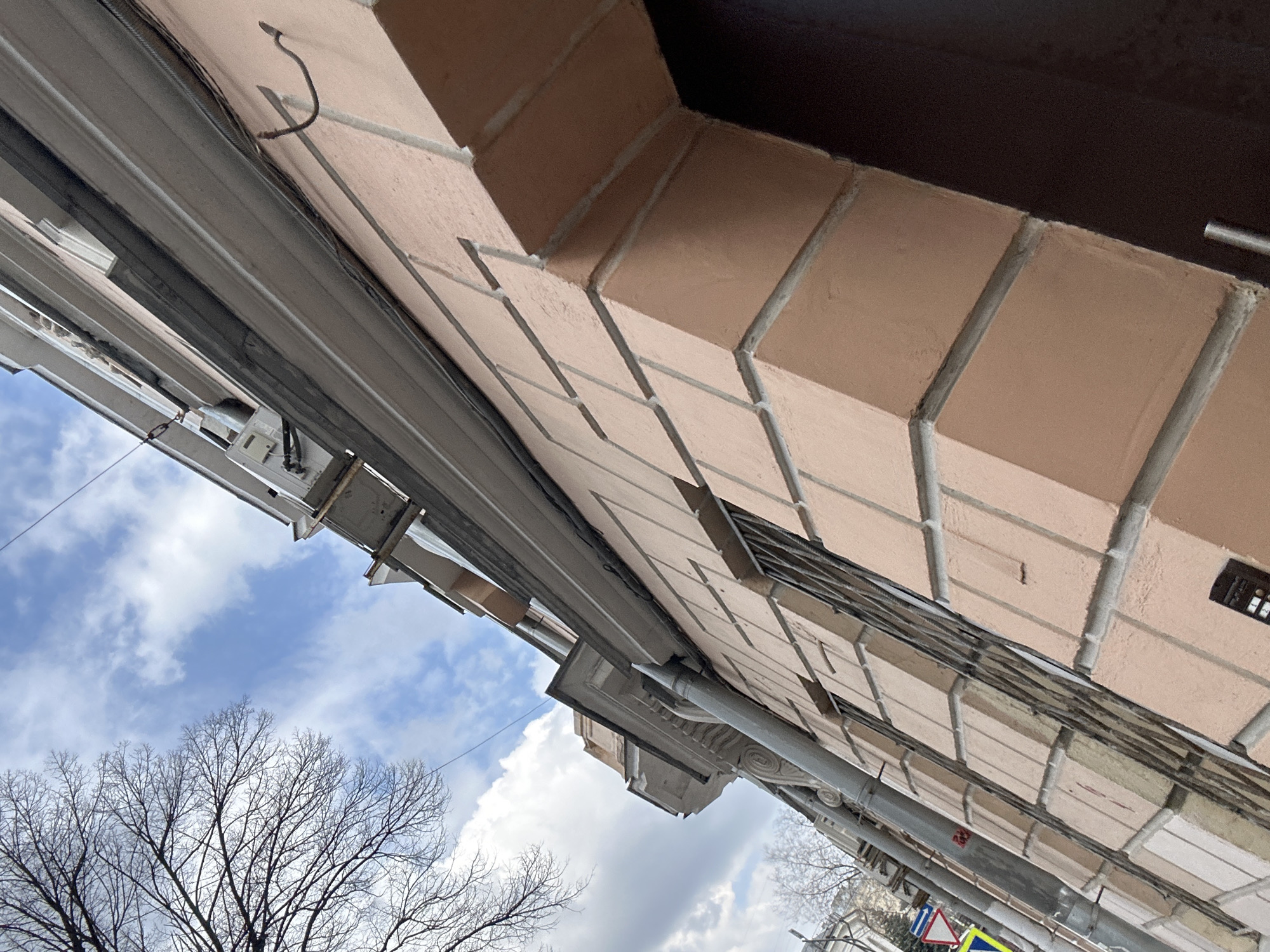 The image shows a low-angle view of a building facade, the sky, and tree branches. The building is constructed of brick, painted a light peach color with grey mortar. Various pipes and electrical wiring are visible, running along the facade. The sky is cloudy with patches of blue. The bare branches of trees are visible in the lower left of the frame. A street sign is also partially visible on the lower right. It is likely in the city of Moscow, Russia.