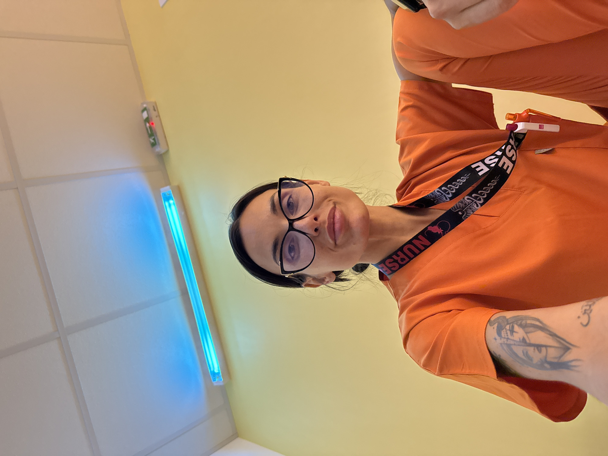 A woman wearing glasses and an orange scrub suit takes a selfie in a room with yellow walls and a white ceiling. A long, blue fluorescent light fixture is visible on the left wall. A "NURSE" lanyard is around her neck, and a tattoo is visible on her left arm. The location is unknown and cannot be determined based on the image.