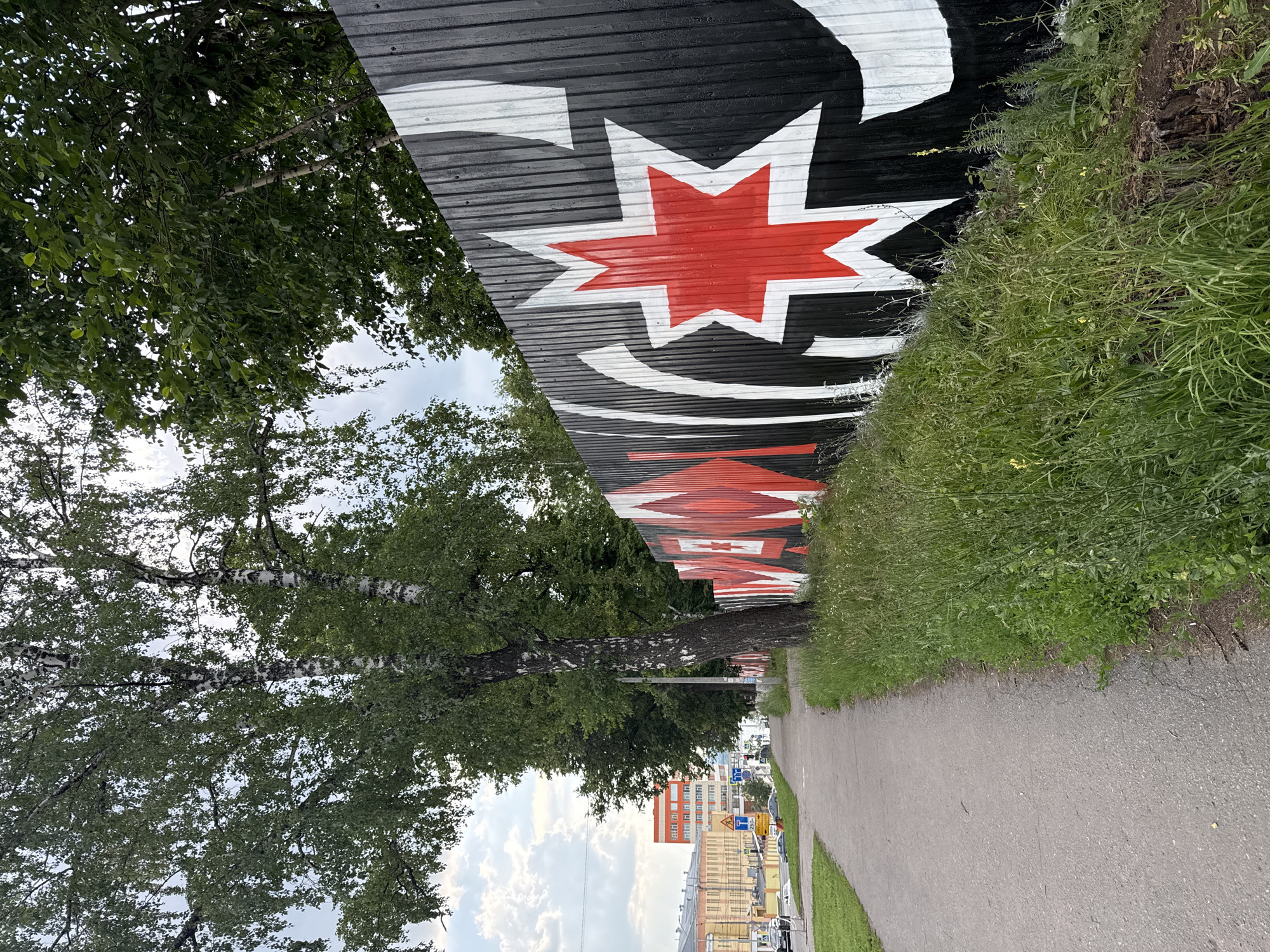 A sidewalk runs alongside a corrugated metal fence decorated with a black, white, and red mural. The mural features geometric shapes and a prominent red star with white outlines. Tall green trees, including birches with white bark, border the fence. In the background, the cityscape of Izhevsk, Russia, is visible, with buildings of various heights and colors lining a street. Cars are present on the street, and traffic lights can be seen. The sky is cloudy. The overall scene depicts an urban environment with artistic elements and natural surroundings.