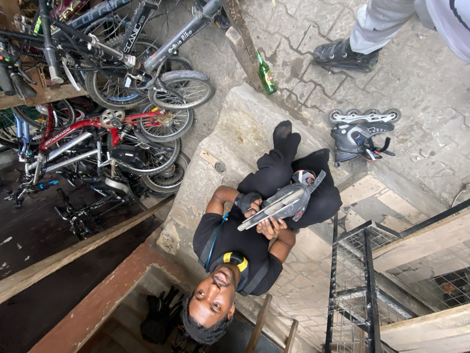 A man in black clothing and black socks sits on a concrete step, holding an inline skate. He is wearing a backpack with yellow accents. Near him are a pair of inline skates, and a green bottle. A person wearing grey pants and black shoes stands nearby. To the left, a cluttered pile of bicycles is visible. The location is in Lagos, Nigeria. A black metal shelving unit is in the background.