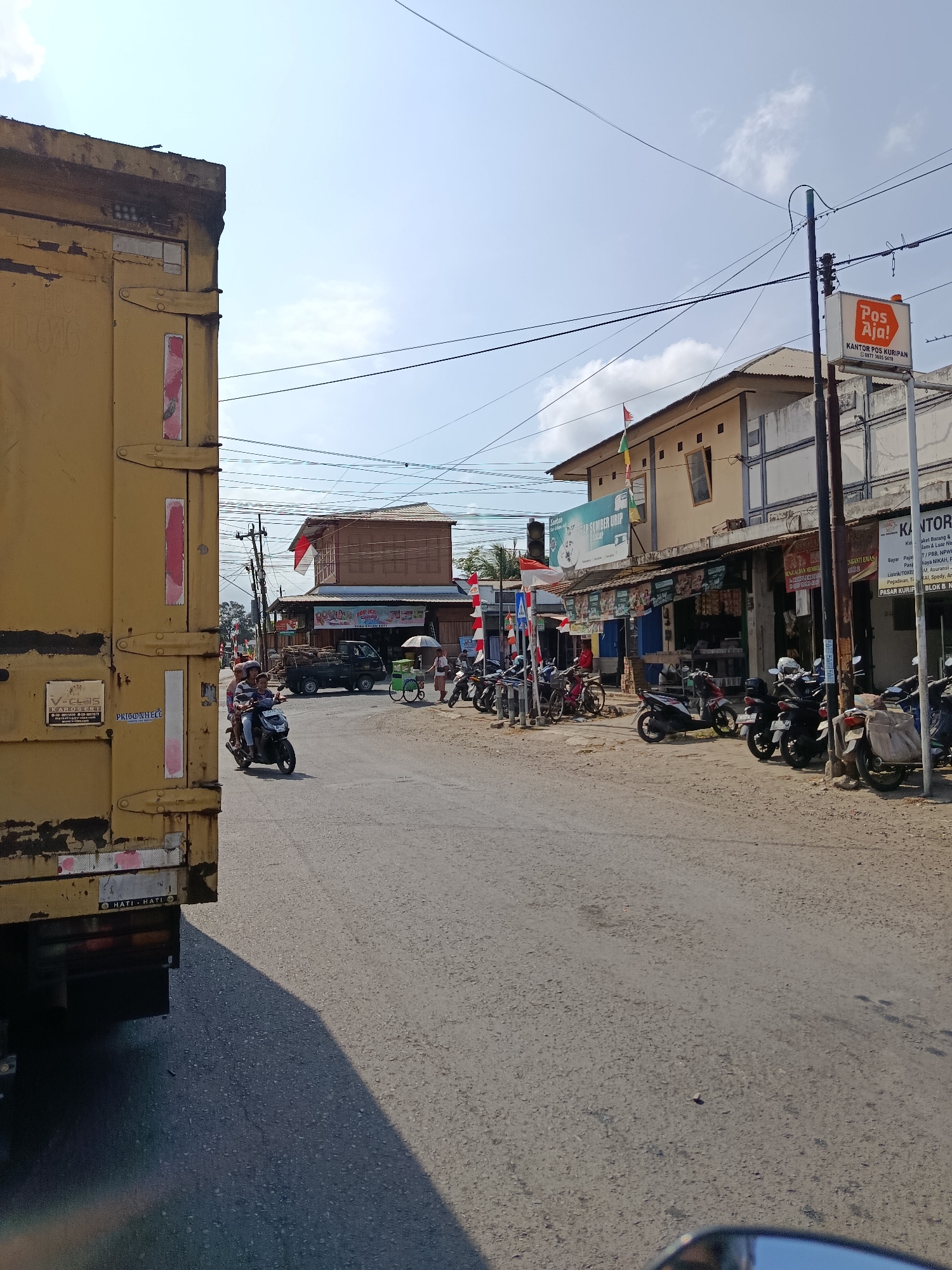 The image depicts a street scene in Kuripan, Indonesia, likely during daylight hours. A large yellow truck occupies the left side of the frame, its rear section visible. The road surface appears to be asphalt, with some patches and debris.
On the right side of the street, a row of buildings lines the road. These buildings appear to be shops or small businesses, with awnings and signage visible. One sign prominently displays "Pos Aja! KANTOR POS KURIPAN". Several motorcycles are parked in front of these establishments.
Several individuals are present in the scene. Two people are riding on a motorcycle in the middle of the road, heading away from the viewer. Another person is walking near the shops, pushing a cart with an umbrella. Other individuals are partially visible in the background, near the buildings and parked vehicles.
The sky is visible in the upper portion of the image, appearing partly cloudy. Power lines are strung across the sky, supported by utility poles. The overall impression is a typical commercial street in a town or city in Indonesia.