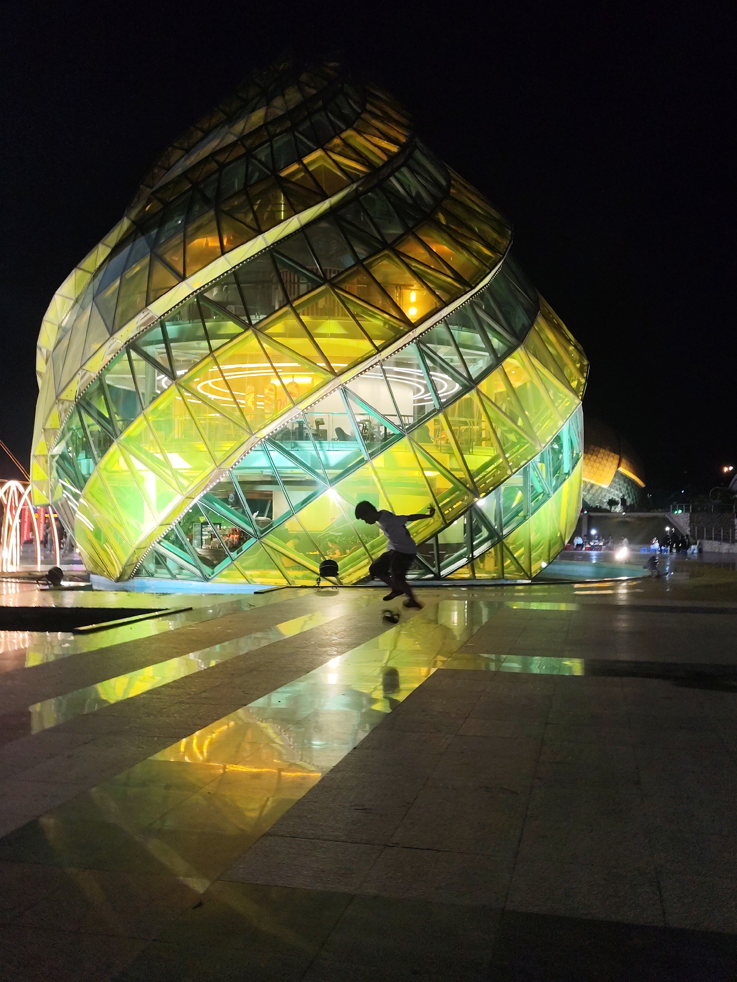 A medium, long shot taken at night captures a person skateboarding in front of the geometrically-designed glass building, the Dalat Market. The building is illuminated with yellow and green light. The plaza is tiled and reflective, reflecting the bright light of the building. A lone skateboarder is in the midst of an ollie, positioned in the center-right portion of the frame. The background is dark, with a few blurred lights visible in the distance.