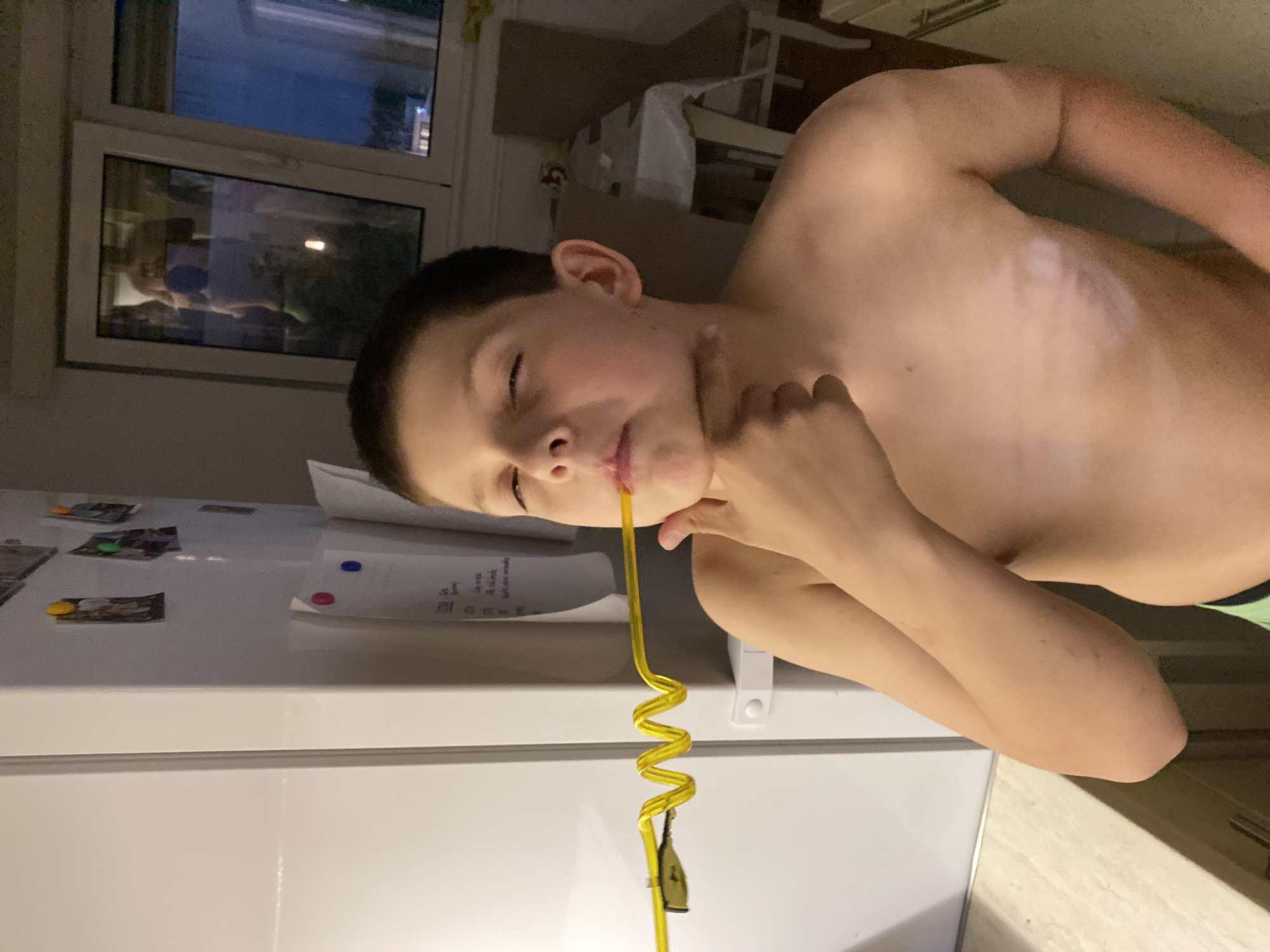 A shirtless boy is drinking from a long, yellow, swirly straw. The boy is leaning over a white refrigerator, which has magnets and papers attached to it. There is a window and a chair visible in the background. There is no information indicating this photo was taken in Budva, Montenegro.