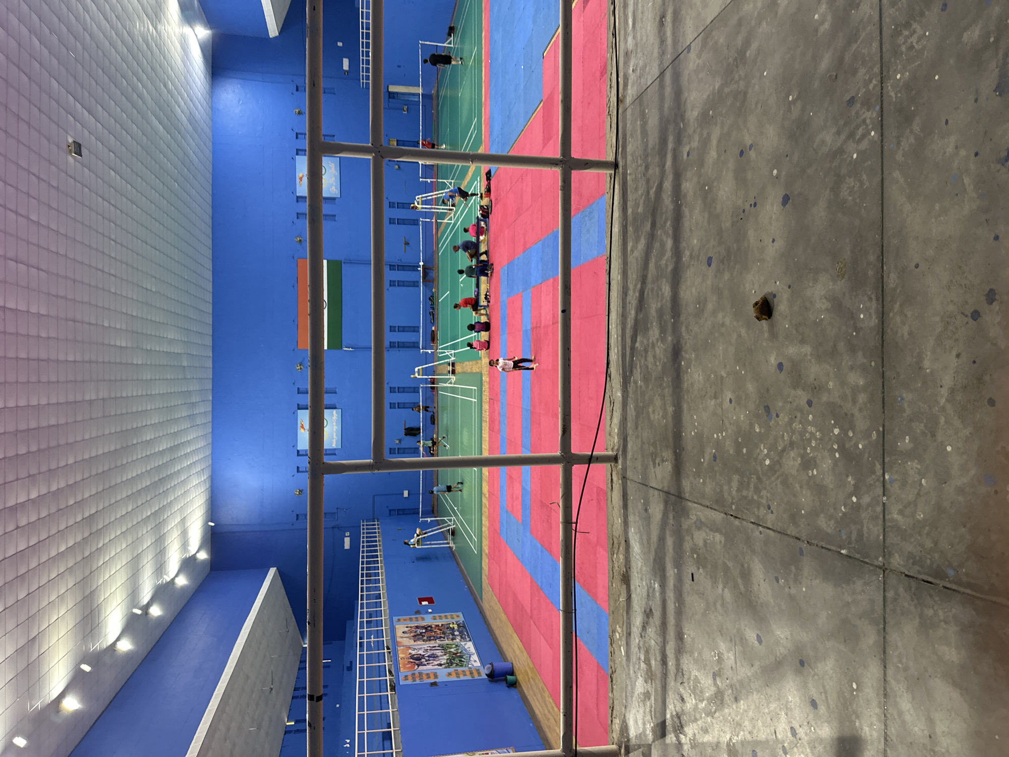 An indoor sports facility with a blue wall and a white ceiling with recessed lighting is visible. The floor is made of red and blue mats, and there are several badminton courts marked out with green flooring. People are scattered across the courts, with some playing badminton and others sitting and watching. A young girl in a pink shirt and white shorts is standing on the red mat. A brown object, possibly a piece of debris, rests on the concrete floor in the foreground. A railing separates the viewer from the sports area.