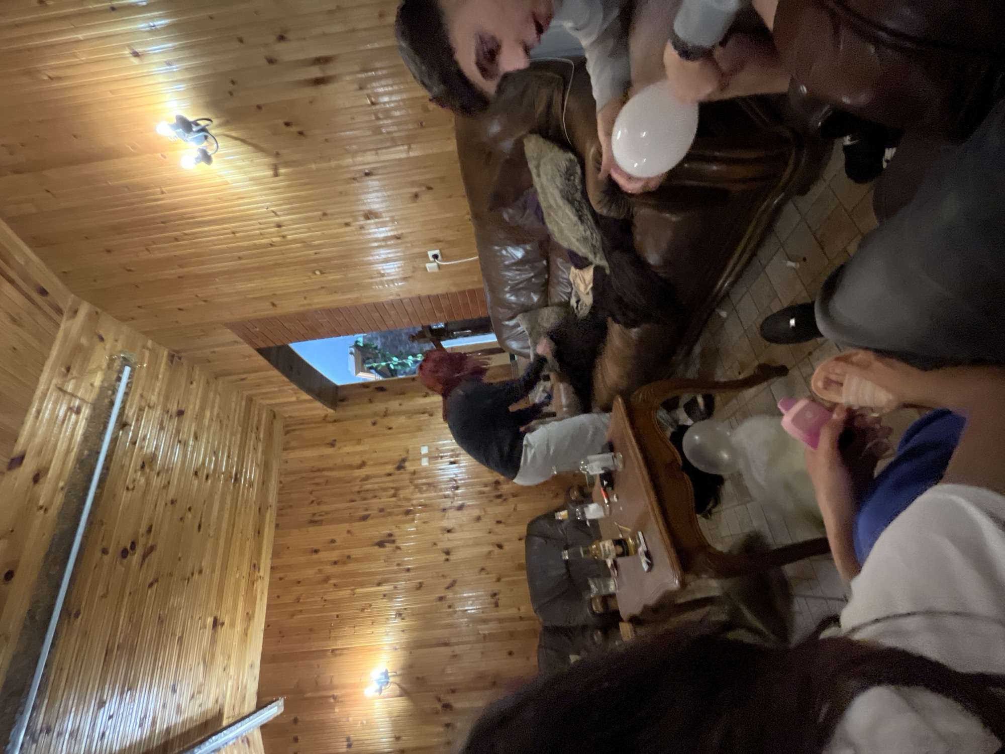 The image shows an interior scene of a room with wood-paneled walls and ceilings. A group of people is present. One person is seated on a brown leather sofa holding a white balloon. Another person with red hair is seated at a wooden table with bottles and glasses. Other individuals are partially visible in the frame. The specific location in Presoka, Bulgaria, cannot be determined.