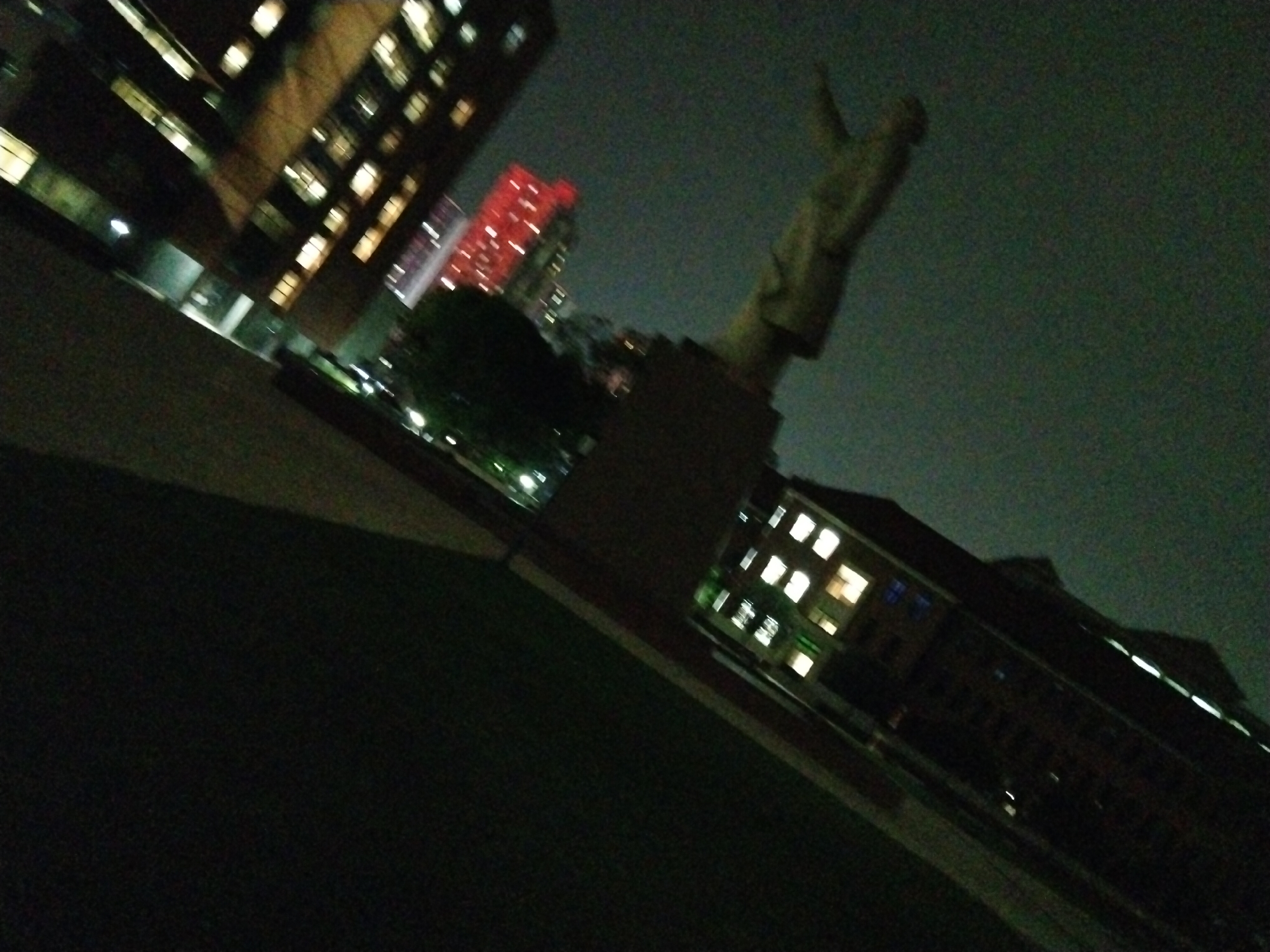 The image shows a nighttime urban scene, likely taken from a low angle. In the foreground, there is a dark area, possibly a grassy lawn or pavement. Several buildings are visible in the background. On the left, a tall building with illuminated windows stands out. Further to the right, there is a red-lit building that appears taller than the first building. In the center of the image, a statue is erected on a pedestal. There are lights on buildings to the right. The photo is angled, making it difficult to determine the exact layout of the area. The lighting in the scene is dim, with artificial lights illuminating the buildings and statue. The image is grainy, suggesting a low-light photography setting. There is nothing in the image to indicate a location.