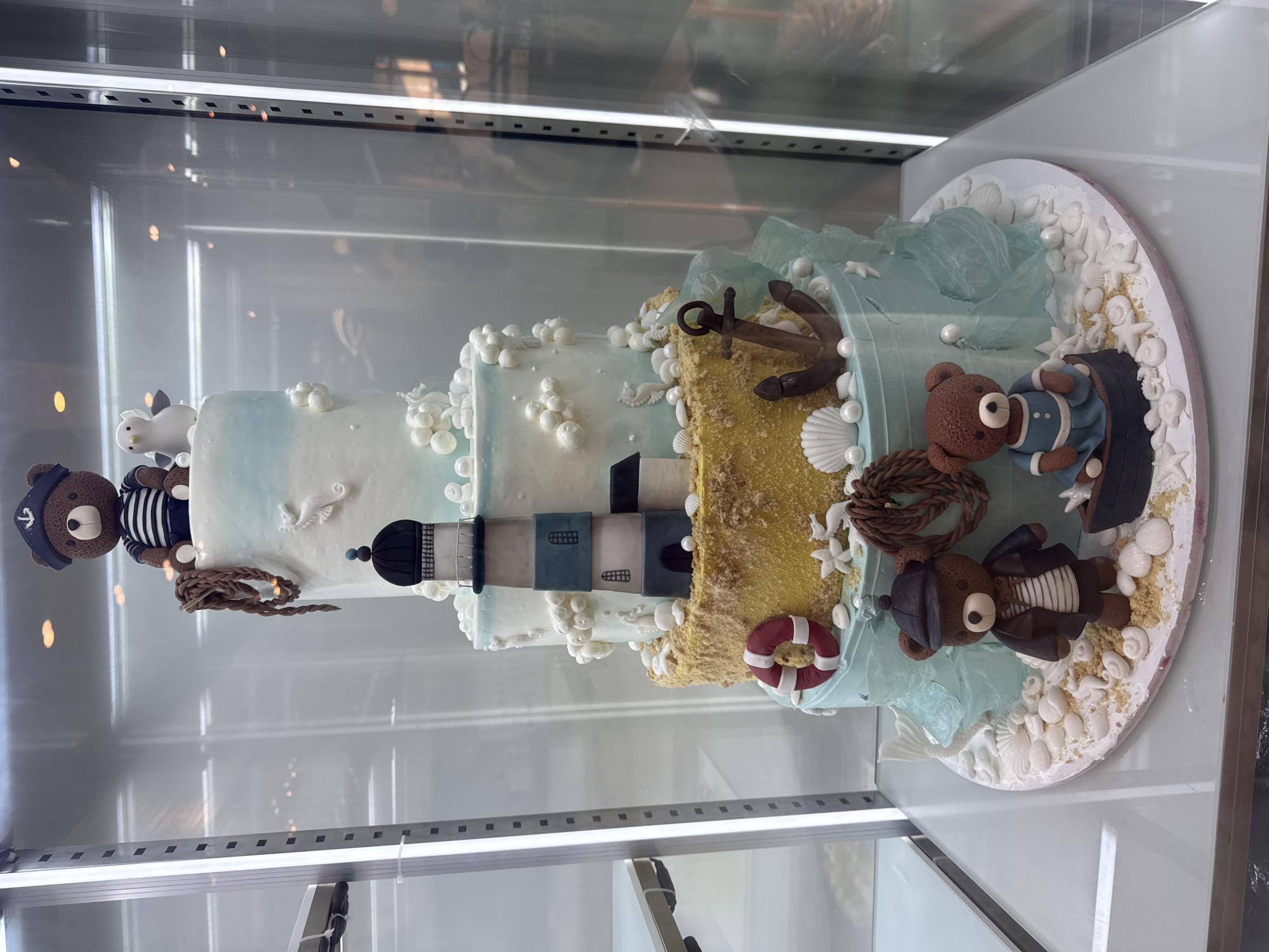 The image depicts a multi-tiered cake displayed inside a glass case, likely within a bakery or confectionery shop. The cake has a nautical theme with elements such as a lighthouse, anchor, life preserver, rope, seashells, edible "sand," and fondant bears. The colors are predominantly white, blue, and beige, with some brown and black accents. The frosting is smooth with applied decorations like pearls and sea life shapes. The background is a blurred reflection within the glass case. There is no evidence of the city or location; it is not possible to determine this from the provided image.