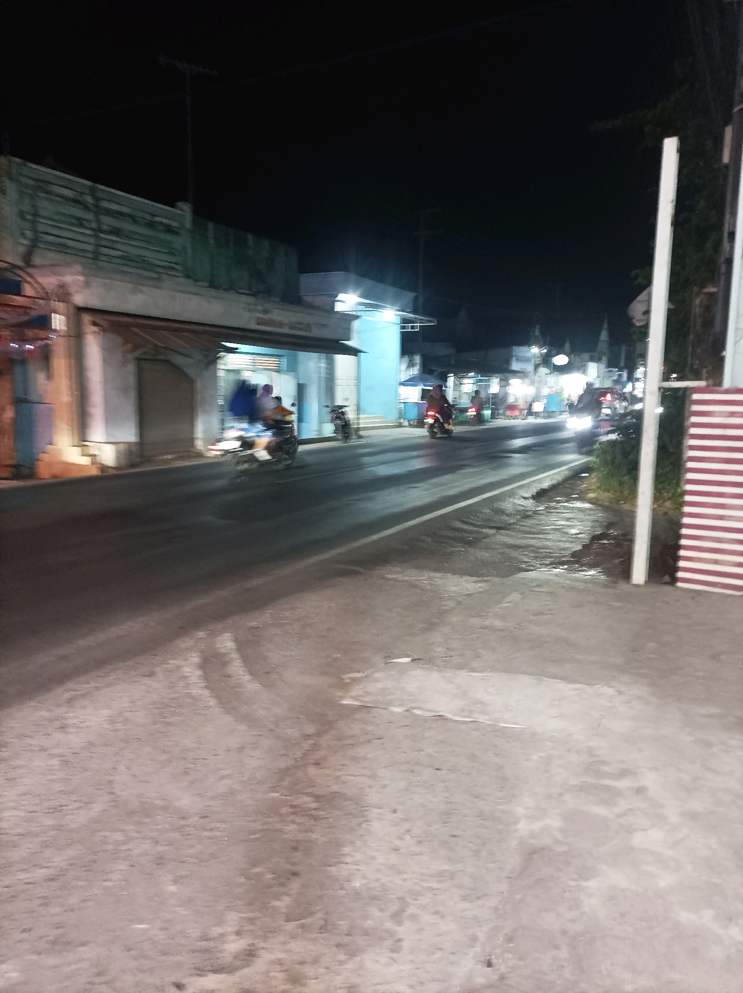 A nighttime street scene in Blimbing, Indonesia, shows a road with several motorcycles in motion, indicating traffic. On the left side of the road, buildings with closed storefronts are visible, some with illuminated signs. The road surface appears wet, reflecting the lights from the vehicles and buildings, suggesting recent rain or a damp environment. A white pole with some greenery next to it stands on the right side of the frame, and a structure with horizontal red and white stripes is partially visible in the far right. The sky is dark, indicating it is late at night.