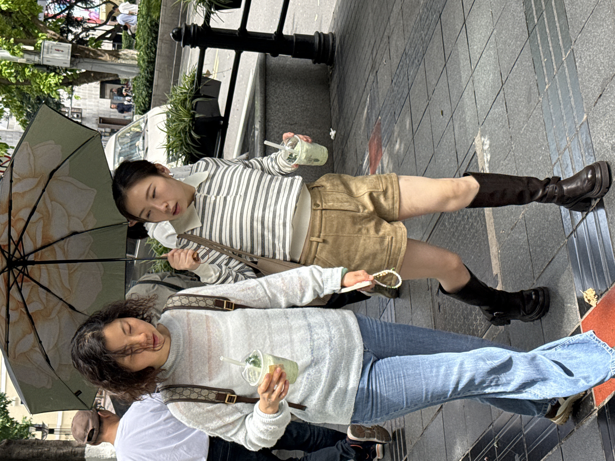 In Yuzhong District, China, two young women are walking on a city street. The first woman wears a striped shirt, beige shorts, knee-high brown boots, and carries a beige purse. She holds a drink in her hand and an umbrella protects them from the sun. The second woman wears a light gray sweater, blue jeans, and sneakers. She also holds a drink and is partly covered by the umbrella. A man in a white shirt walks alongside them. The street is paved and has metal railings. Trees and buildings are visible in the background.