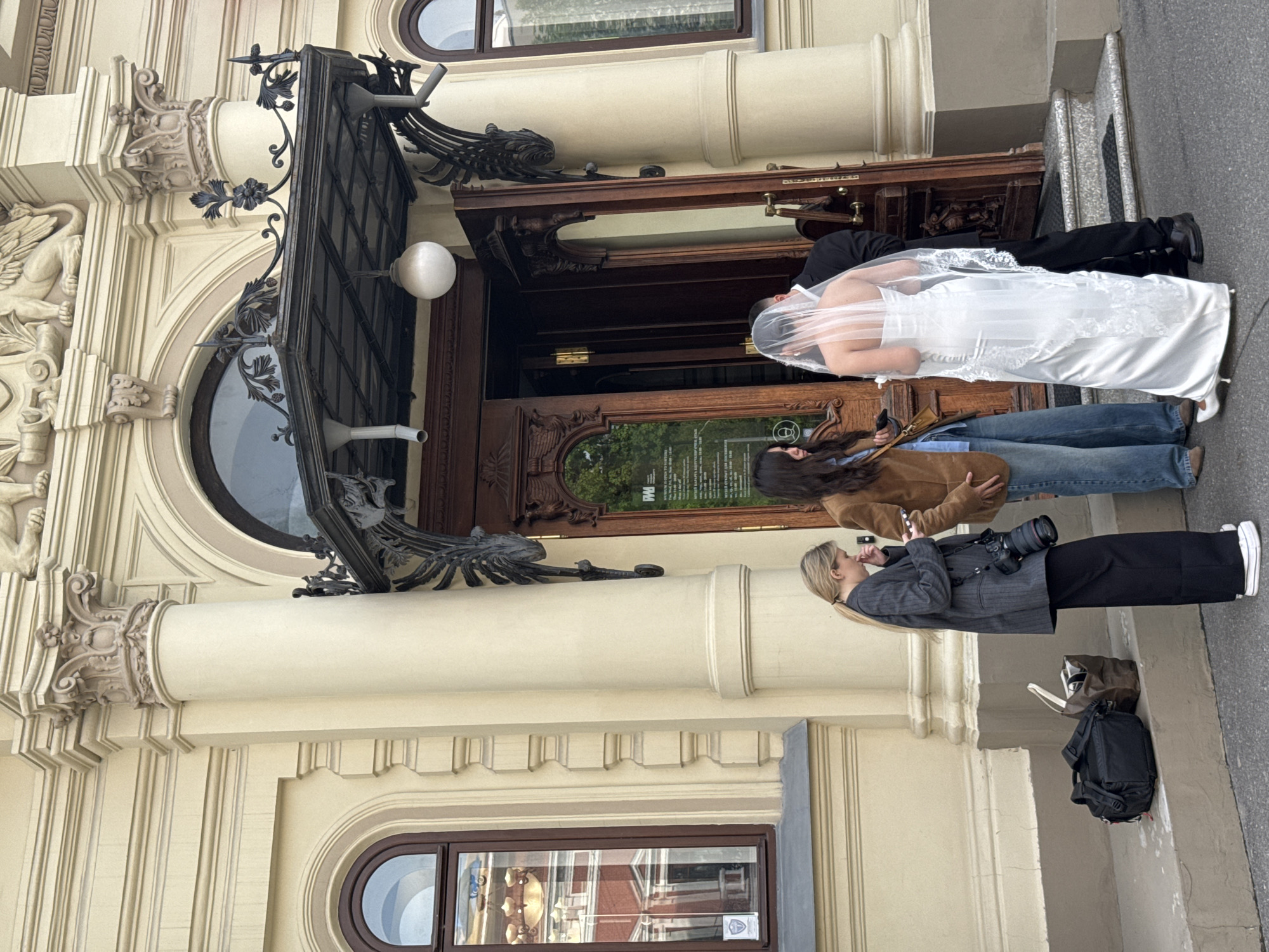 In Kazan, Russia, a wedding couple in formal attire (white dress with veil, black suit) stands in the entrance of a beige, ornately decorated building with a large, dark wood door under a dark metal awning. A woman with blonde hair and wearing a gray blazer and black pants is taking a photo of the couple with a professional camera. Another woman with long brown hair and wearing a brown jacket and jeans is standing nearby. A black backpack and brown bag are on the ground near the photographer. The building features detailed architectural elements including columns, arched windows, and sculptural relief.