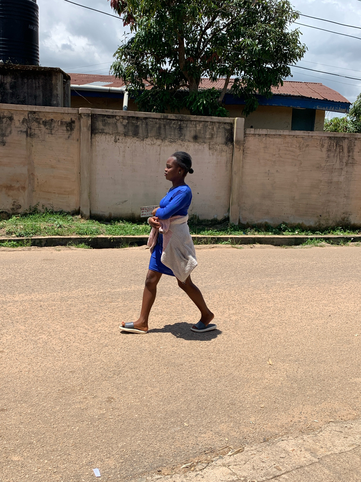 A full shot, taken outdoors in Jos, Nigeria, captures a young Nigerian woman walking on a paved road under a cloudy sky. She is wearing a bright blue dress and a light-colored shawl draped over her shoulders. Her hair is styled in a bun. She is wearing slip-on sandals. The road is empty except for a few small pieces of debris. In the background, a tall concrete wall with a weathered texture runs horizontally across the frame, separating the road from a patch of green grass and some trees. Beyond the wall, a building with a corrugated metal roof and a blue trim is visible, partially obscured by a large tree. A black water tank is also visible on the left side of the frame. The overall scene depicts a casual street view in Nigeria.