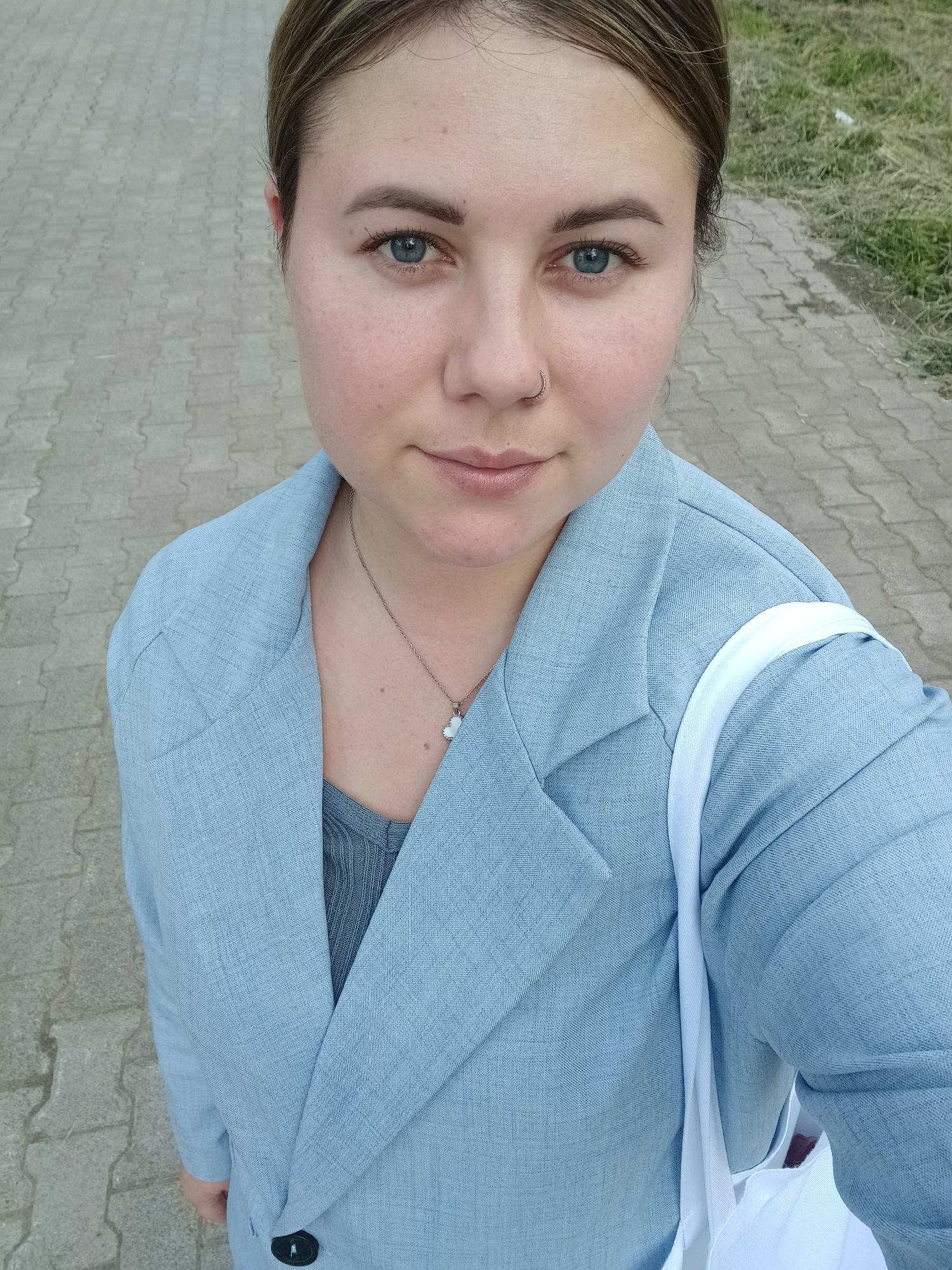 This is a close-up, high-angle selfie of a woman from the chest up, taken outdoors. The woman has fair skin, light brown hair pulled back, blue eyes, and a nose ring. She is wearing a light blue blazer over a grey top and a delicate silver necklace with a white heart pendant. A white strap, likely from a bag, is visible over her right shoulder.
The background consists of a paved walkway made of rectangular interlocking pavers, with a strip of dry grass and some discarded white debris to the right. The lighting suggests it is daytime, and the overall atmosphere is casual. The woman's expression is neutral, with a slight, almost imperceptible smile. There is no visible text in the image. The scene appears to be a public outdoor space, possibly a park or a walkway.
