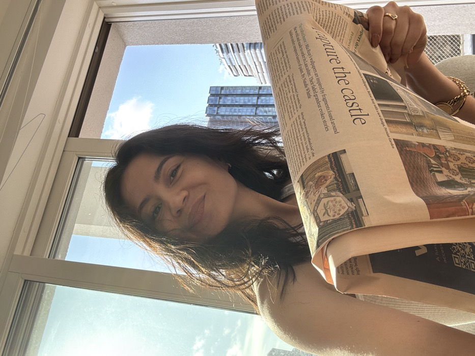 The image depicts an adult female indoors, positioned in front of a large window, smiling directly at the viewer. She has medium-length dark hair and wears a light-colored top. Her left hand, adorned with a gold-colored ring on the fourth finger and a gold-colored chain bracelet, holds a large-format newspaper. The newspaper, light brown in tone, features the headline "Capture the castle," an article about "Mia Perry's wallpapers" by Natalie Whittle, and an illustration of a historical tapestry or crest. A QR code is visible on the lower left portion of the newspaper.
The background through the window reveals a clear blue sky with scattered white clouds and an urban cityscape under bright daylight. Prominently featured on the left is One Blackfriars, a tall, dark, reflective glass skyscraper known as The Vase. To the right of One Blackfriars and further in the distance, another lighter-colored building with a multi-layered or wavy facade is visible.
Based on the identifiable landmarks, specifically One Blackfriars, the scene is located on the north bank of the River Thames, within the **City of London, United Kingdom**, with the view looking south-west towards the South Bank in the borough of Southwark.