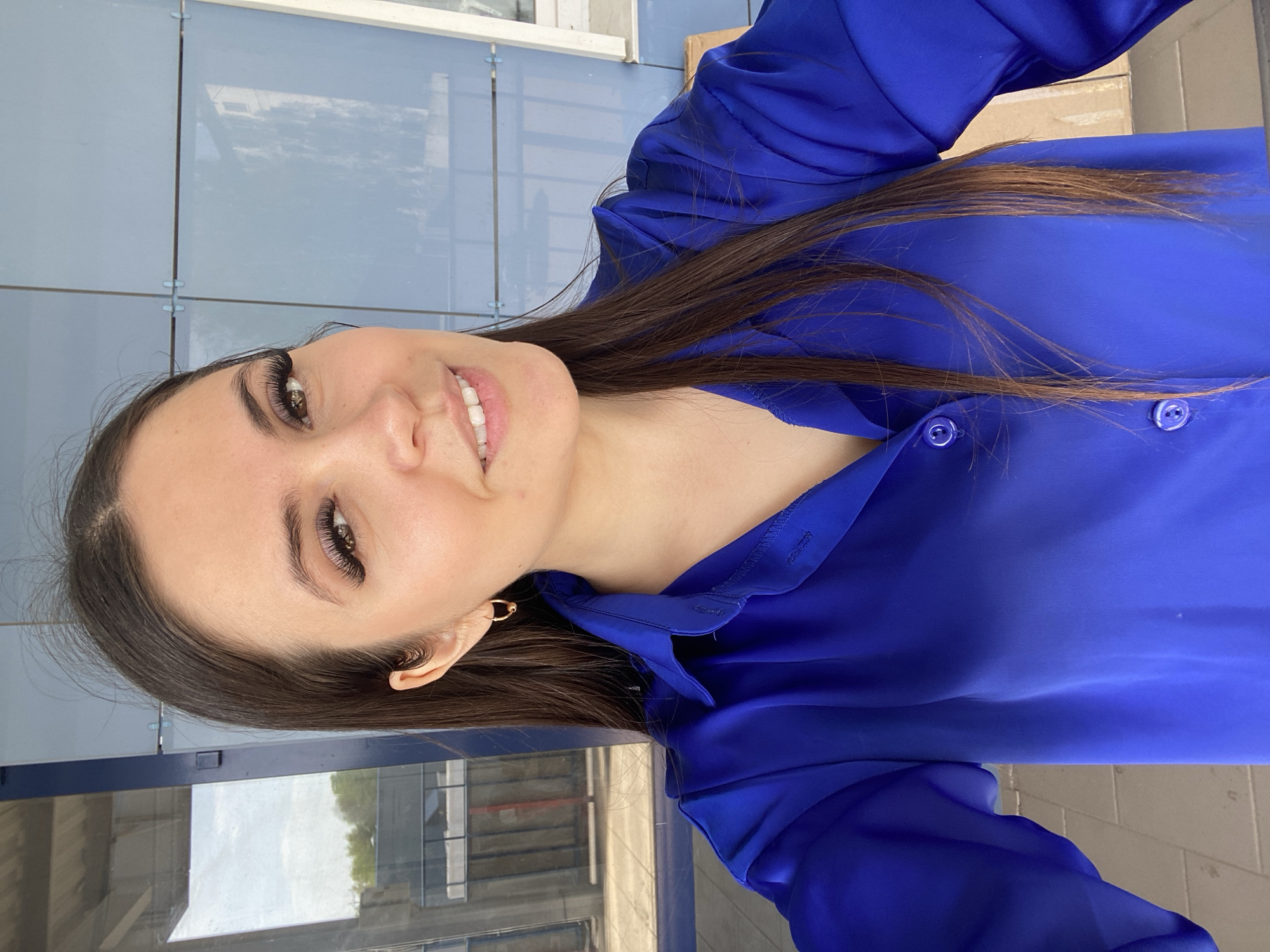 A woman with brown hair, brown eyes, and long eyelashes is looking at the camera and smiling. She is wearing a blue button-down shirt. She has a gold hoop earring in her left ear. The background shows a building with glass windows and a light-colored tiled exterior. The location is the city of Chaikovsky, Russia.