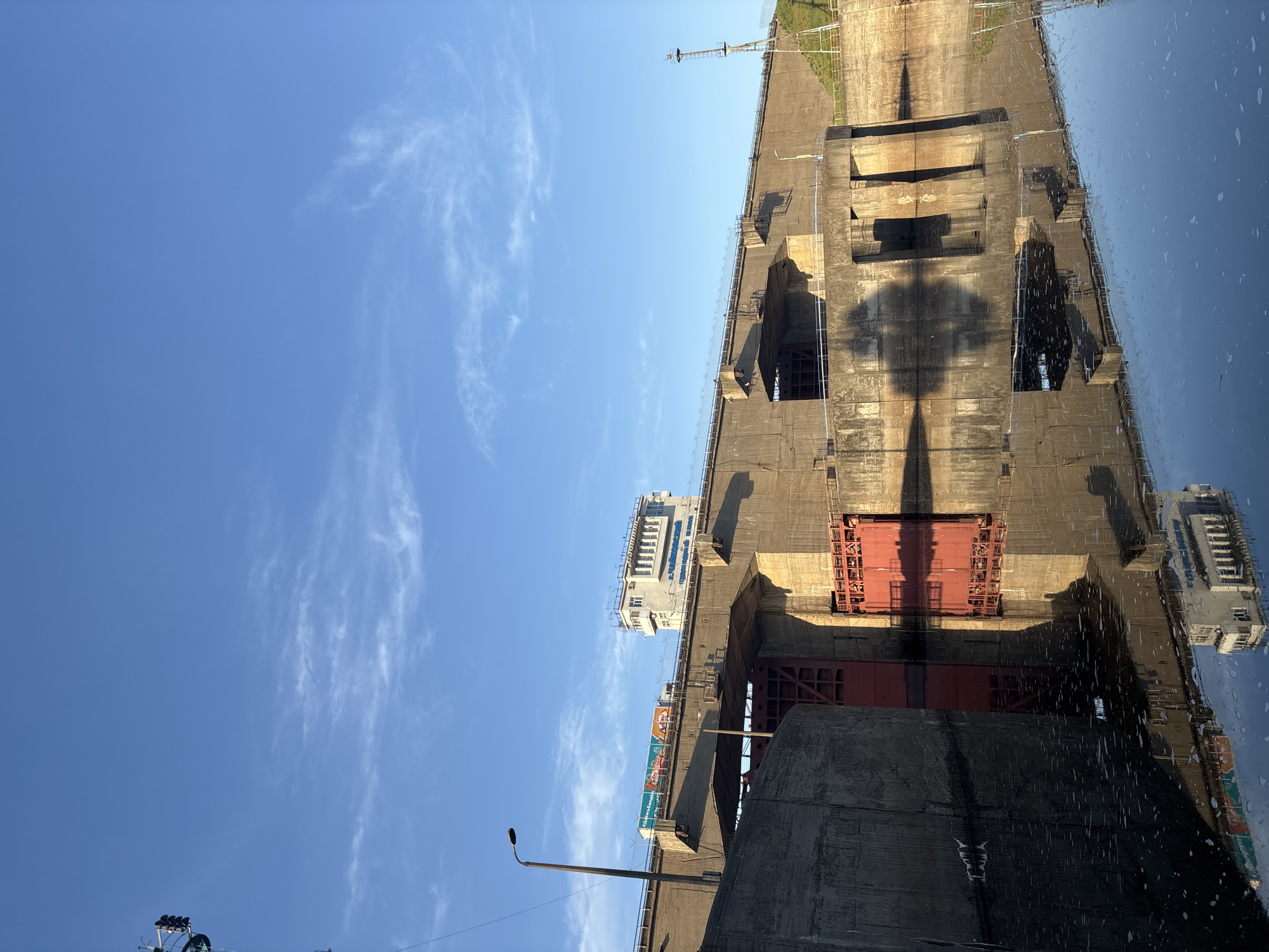 The image shows a view of a hydroelectric dam, possibly the Votkinsk hydroelectric dam located in Chaikovsky, Russia. The dam is constructed of concrete and has a red gated structure. Some parts of the dam and the buildings are reflected in the water below. The sky is blue with some white clouds.