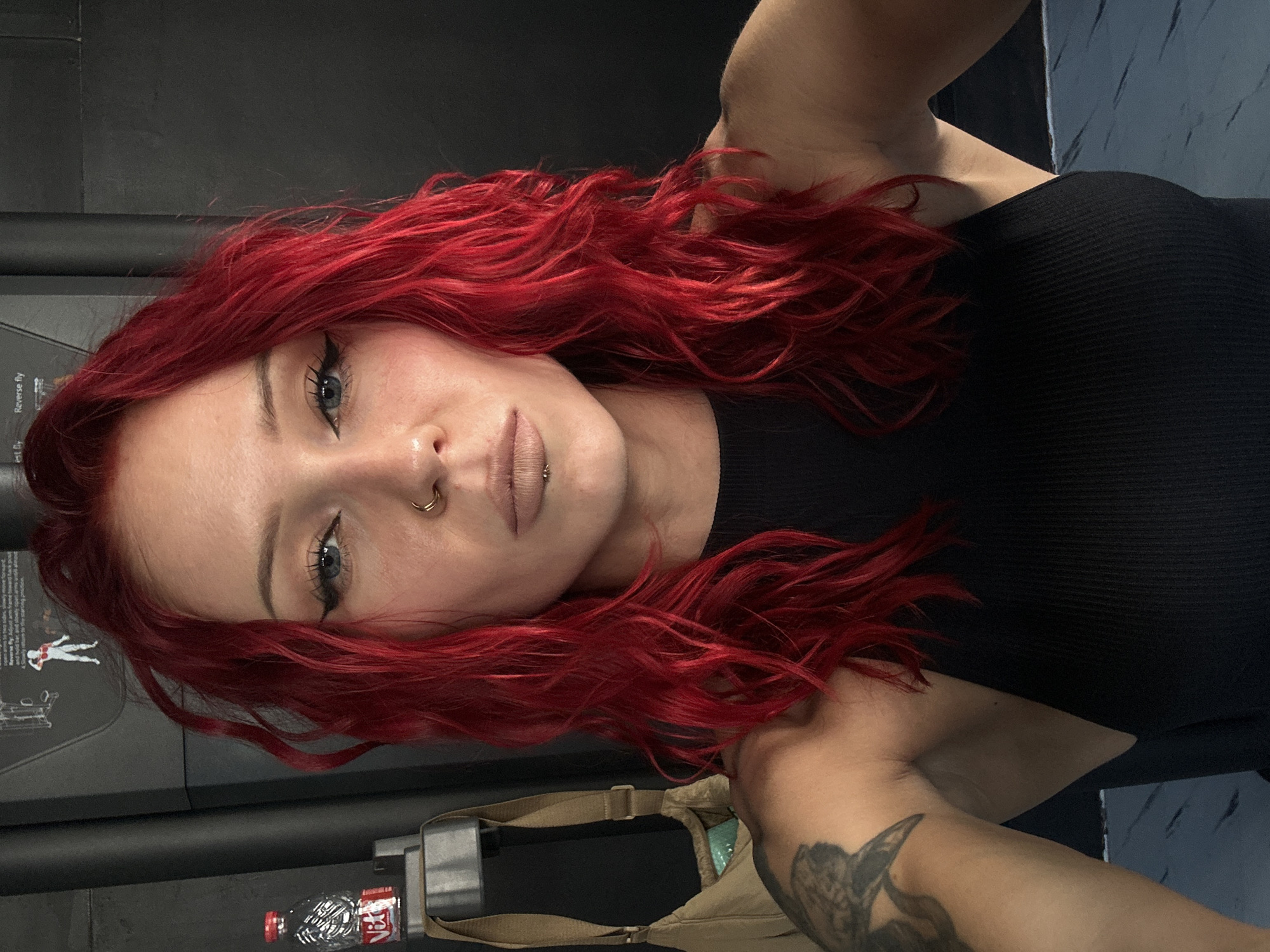 A young woman with red, wavy hair and fair skin is posing for a portrait. She wears a black top and has a nose ring and lip piercings. Her makeup includes winged eyeliner. Visible on her right arm is a dark tattoo. The background appears to be inside a gym, evidenced by the presence of exercise equipment and a water bottle near the bottom of the frame. The location is North Kuta, Indonesia.