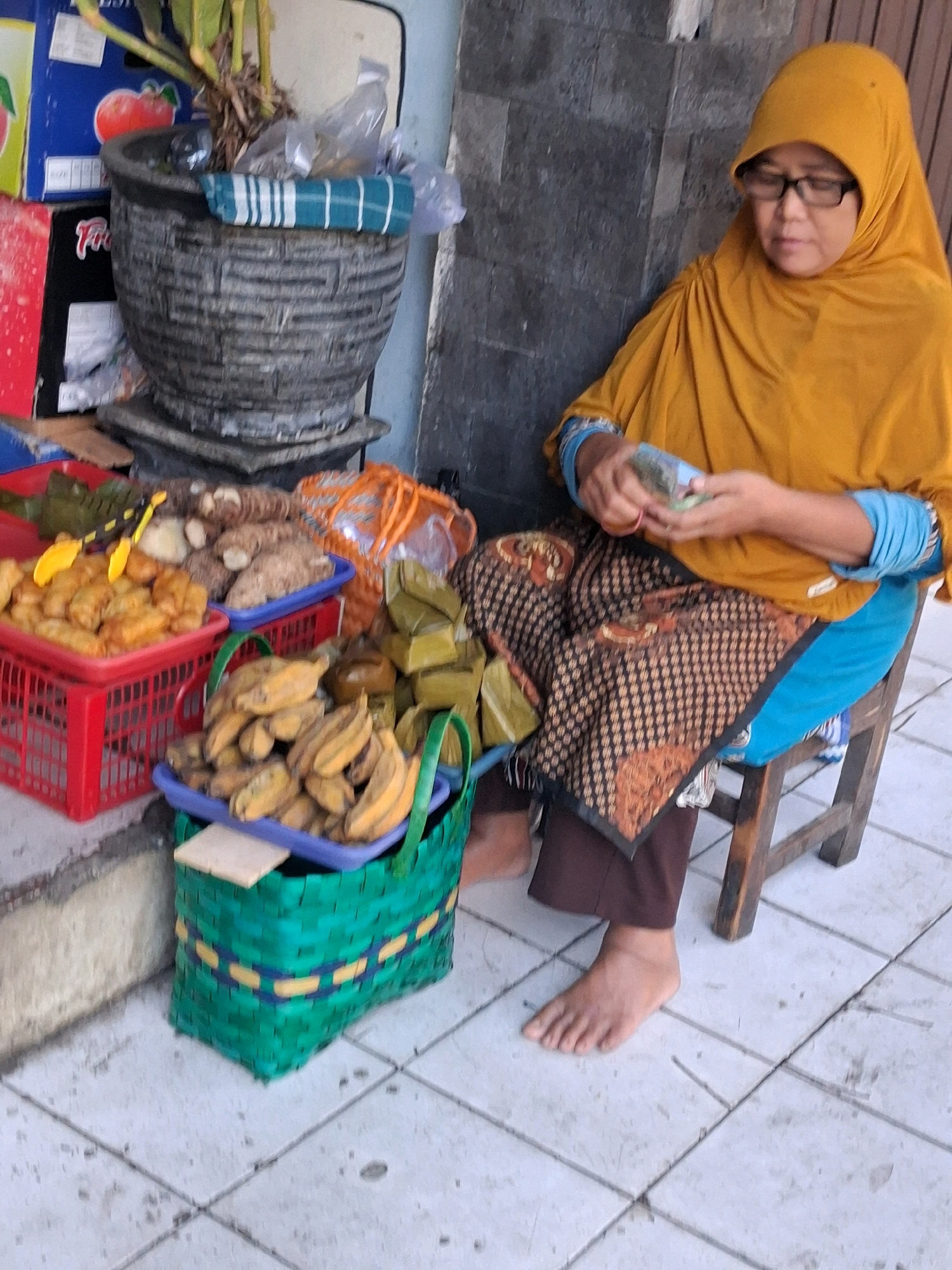 A woman wearing a yellow hijab and glasses is seated on a wooden stool, tending to a street vendor stall in Surakarta, Indonesia. She is holding a small stack of Indonesian Rupiah currency in her hands. The stall displays a variety of food items, including bananas in a green woven basket, fried snacks in a red basket, and what appear to be root vegetables and packaged food items. A large potted plant sits behind the stall. The ground is tiled.