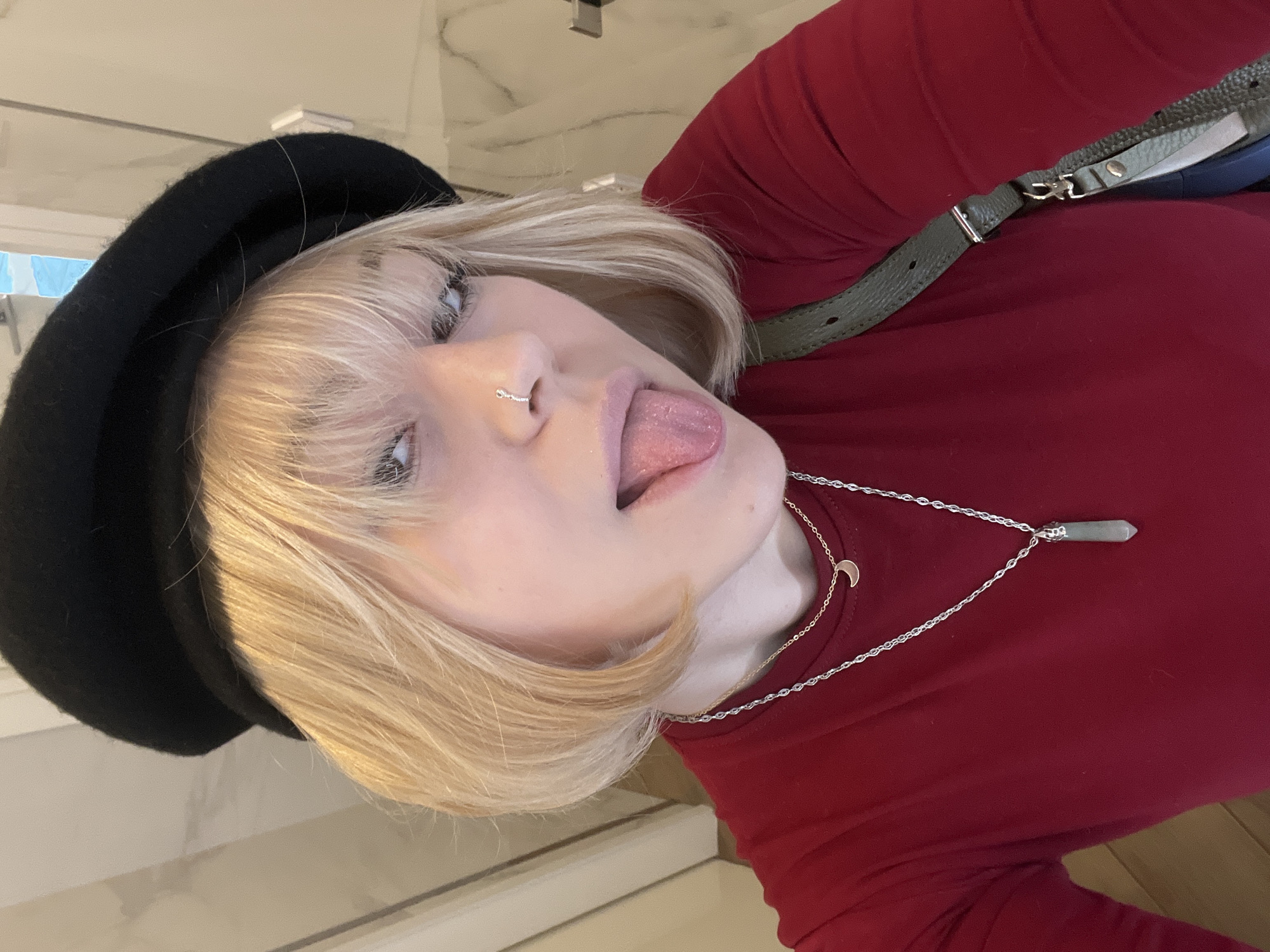 A fair-skinned person with blonde hair, wearing a black beret and a red, long-sleeved shirt, is sticking their tongue out in a close-up selfie. They have a nose ring and are wearing two necklaces, one with a crescent moon pendant and the other with a crystal pendant. A green bag strap is visible over their shoulder. The background is slightly blurred and features a neutral-toned wall. There is no information present in the image that allows to deduce the location in the city of Odesa, Ukraine.