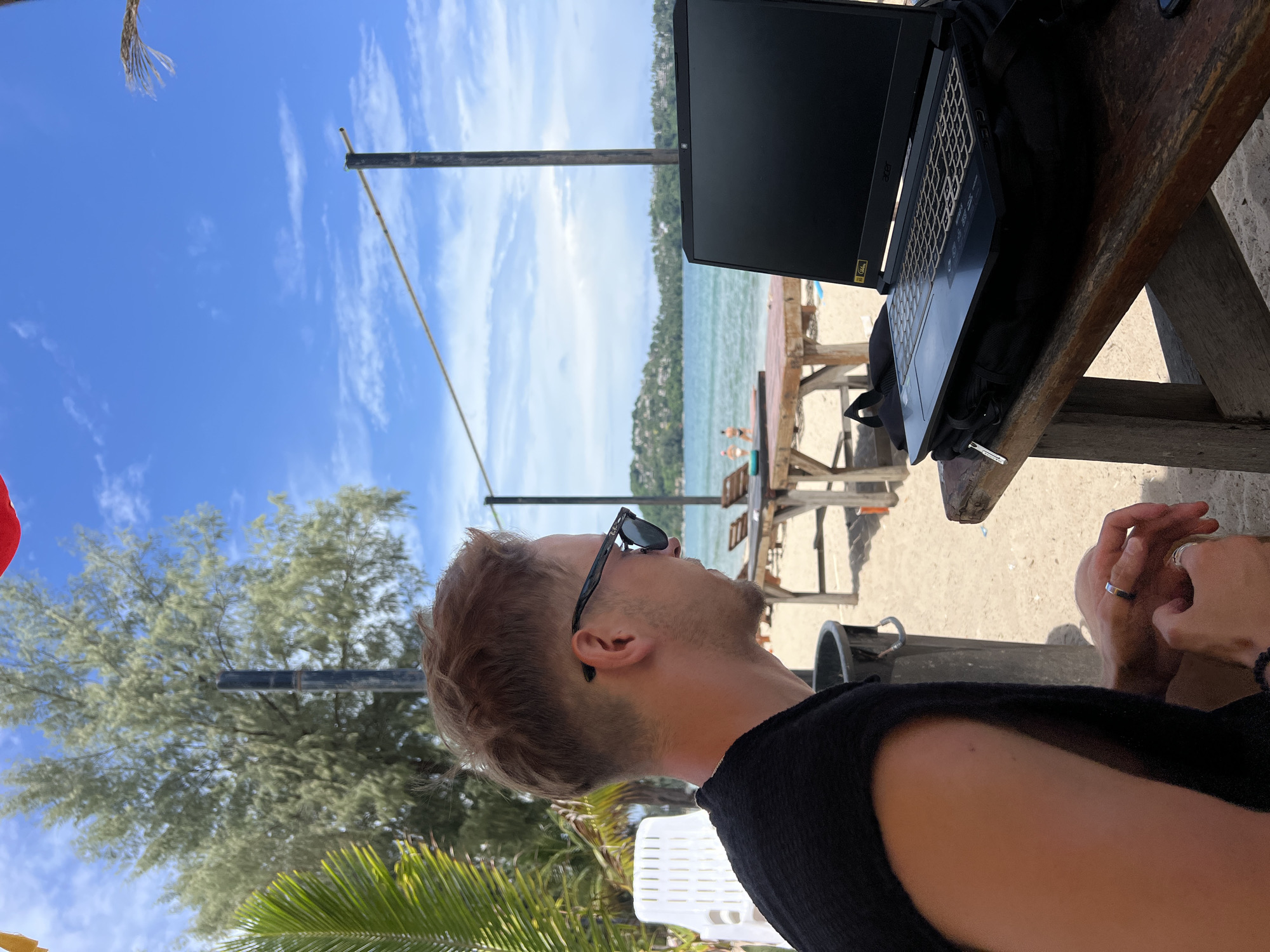 A man with short, blondish-brown hair and sunglasses is seen from the side, sitting at a wooden table on a sandy beach. He is wearing a black sleeveless shirt and appears to be working on a laptop. The laptop is open and placed on top of a black backpack. In the background, there is a body of water with a few people visible, a line of trees, and a blue sky with scattered clouds. The overall scene suggests a relaxed, outdoor work or leisure setting.
