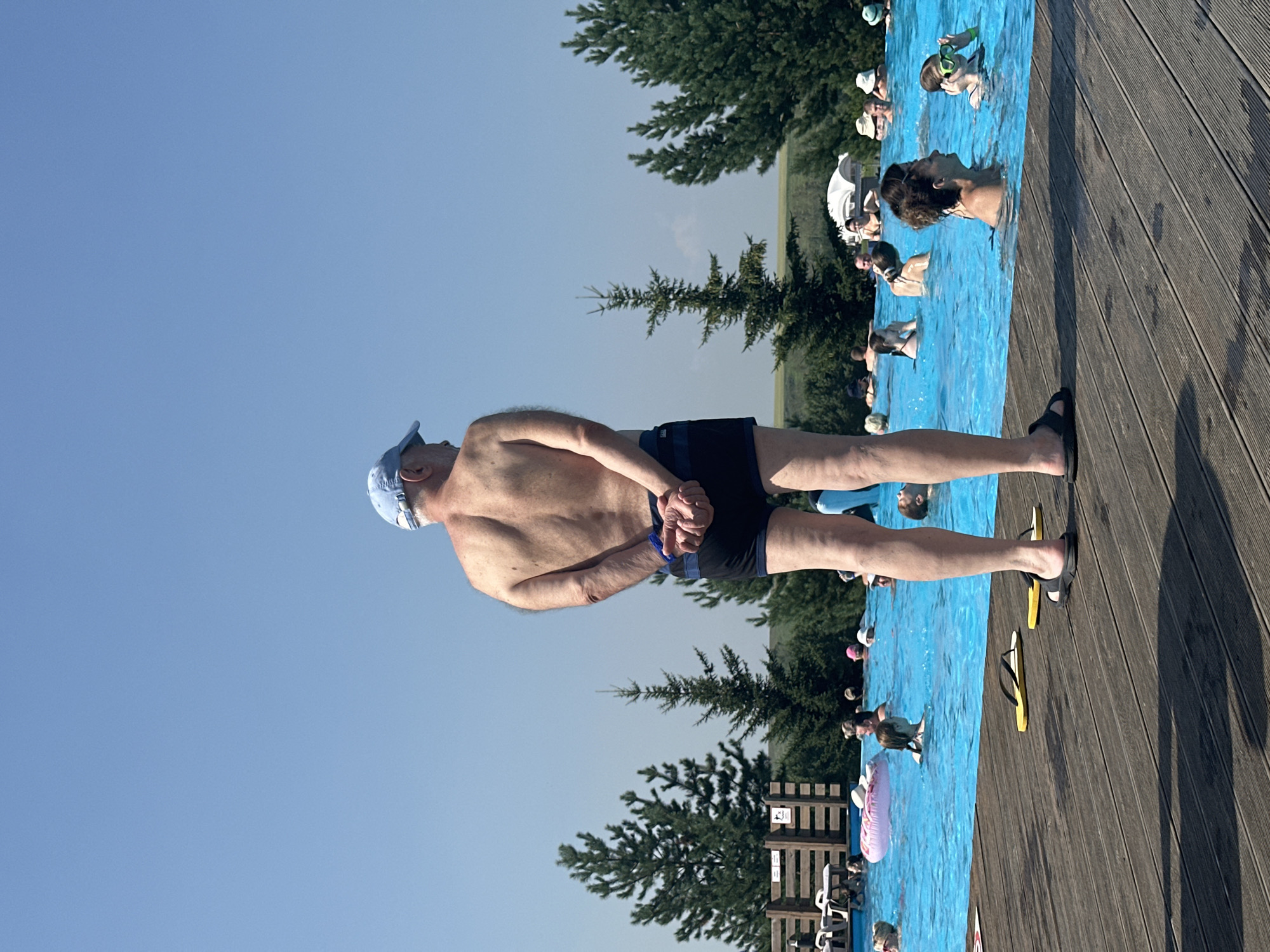 An outdoor swimming pool scene is depicted on a sunny day. The focus is on a man standing on a wooden deck, facing away from the camera, wearing a blue cap and dark swim trunks. His hands are clasped behind his back. The pool is filled with bright blue water, and several people are visible swimming and relaxing in it. Trees, including pine trees, line the edge of the pool, with a clear blue sky overhead. The overall impression is a lively recreational setting.