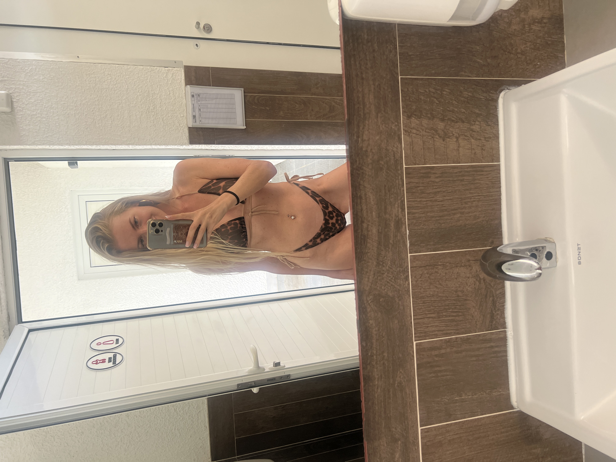 A woman with long blond hair is taking a selfie in a bathroom. She is wearing a leopard print bikini. The bathroom has brown tile walls and a white sink. There is a sign on the door with symbols of a toilet and a shower. The location is likely in Budva, Montenegro, based on the user's prompt.