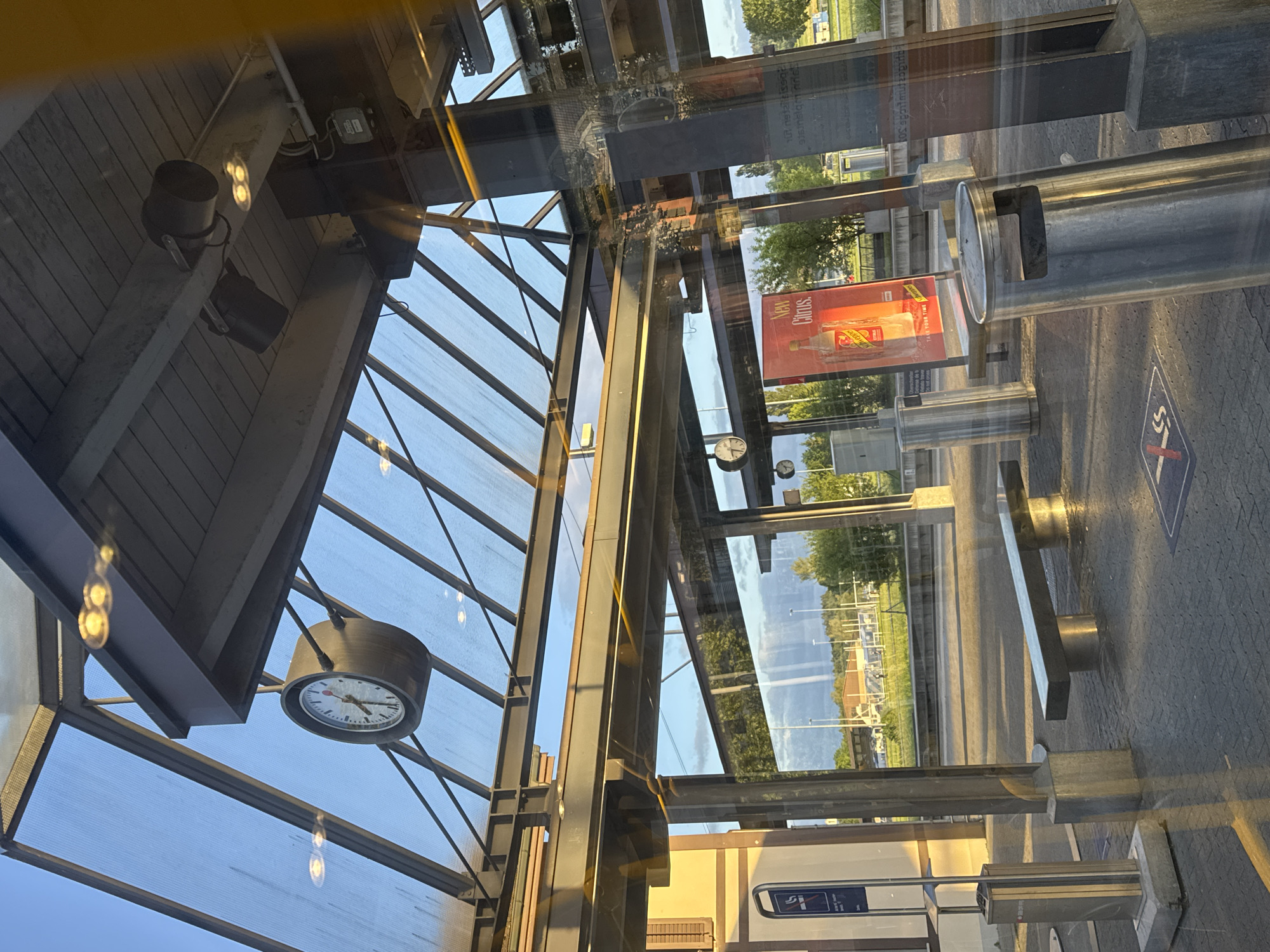 This image captures the interior of a modern building, likely a public transport stop or waiting area, looking out through large, reflective glass panels. The scene is brightly lit by natural light, suggesting a clear day.
The interior architecture features a ceiling constructed with horizontal wooden slats and robust metal beams. Several dark spotlights are mounted on these beams, and light reflections are visible on the glass. A prominent round clock with a dark frame, white face, and black hands hangs from the ceiling, displaying the time as approximately 10:10. More metal structural elements and glass sections complete the indoor framework.
Through the extensive glass, a vibrant outdoor environment is visible. Green trees and grass form a backdrop, indicating a natural setting beyond the immediate vicinity. A large, rectangular billboard, primarily red, is clearly visible, advertising "New Citrus. Schweppes." with smaller, less distinct text also present. The ground outside appears paved, with various metallic public amenities including cylindrical bins or ashtrays and a rectangular bench. A blue square sign on the paved ground displays a white circle with a crossed-out cigarette, signifying a "no smoking" zone. Another blue sign with a smoking symbol is affixed to a vertical surface, possibly indicating an ashtray.
The glass panels exhibit strong reflections, superimposing elements of the interior architecture onto the exterior view and creating a layered, somewhat abstract visual effect. The overall impression is of a calm, static scene under bright sunlight, with no people present. The time of day appears to be late morning, given the position of the sun and the clock reading. The context indicates the location is Fehraltorf, Switzerland.